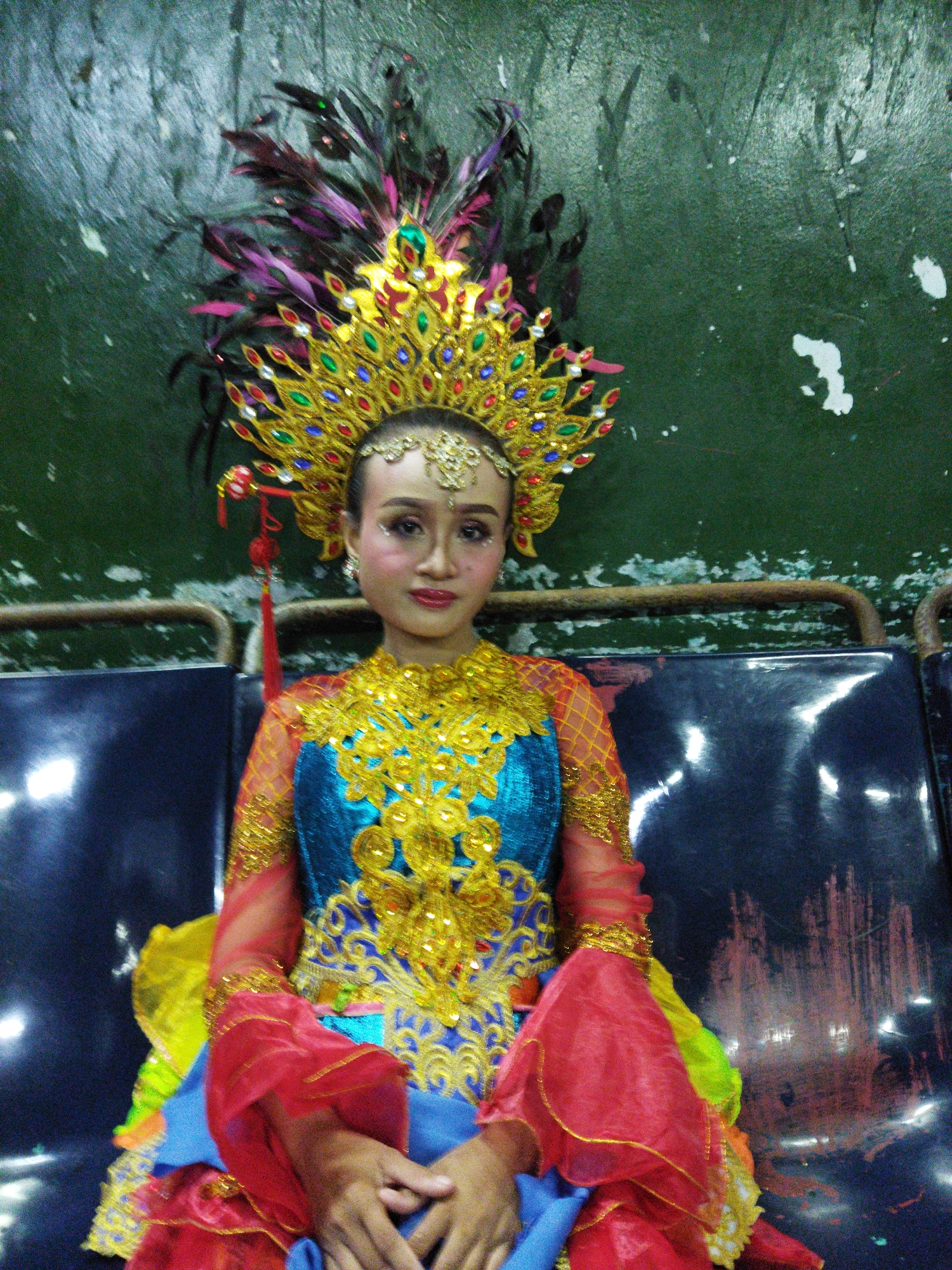 A young female dancer, adorned in a vibrantly colored, ornate costume and headdress, is seated on a dark blue bench, possibly in an indoor venue. The costume features a turquoise and gold bodice with intricate embellishments, sheer red sleeves with gold trim, and a layered skirt of blue, red, and yellow fabric. Her headdress is a large, golden crown adorned with colorful jewels and feathers, with a red tassel hanging to the side. She has dramatic makeup with glitter accents on her eyes and cheeks. The background is a textured, dark green wall with white splotches. The bench appears to be part of a row of similar seating. While the exact location in Semarang, Indonesia, cannot be definitively determined from the image alone, such performances are common in cultural centers, community halls, or event spaces within the city.