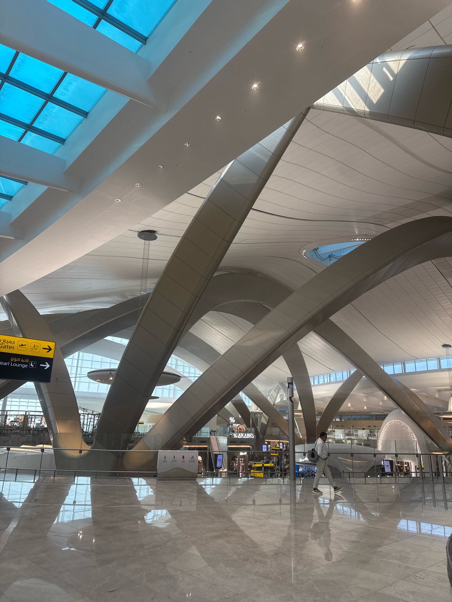 This is a photograph of the interior of an airport terminal. The architecture is modern and striking, with large, curved beams supporting a translucent blue ceiling made of what appears to be glass panels. The floor is highly polished and reflective, showing the shapes of the ceiling structure.
In the foreground, a yellow directional sign is visible with Arabic and English text. It indicates "Gates" with an airplane icon and an arrow, and "Pearl Lounge" with an arrow.
Further into the terminal, a person is walking from right to left. They are wearing a light-colored jacket and pants, with a bag slung over their shoulder. Other commercial establishments are visible in the background, including shops and potentially food vendors. The overall environment is clean, bright, and spacious, typical of a modern international airport. The location is identified as Al Matar, United Arab Emirates, which is the airport zone of Dubai.