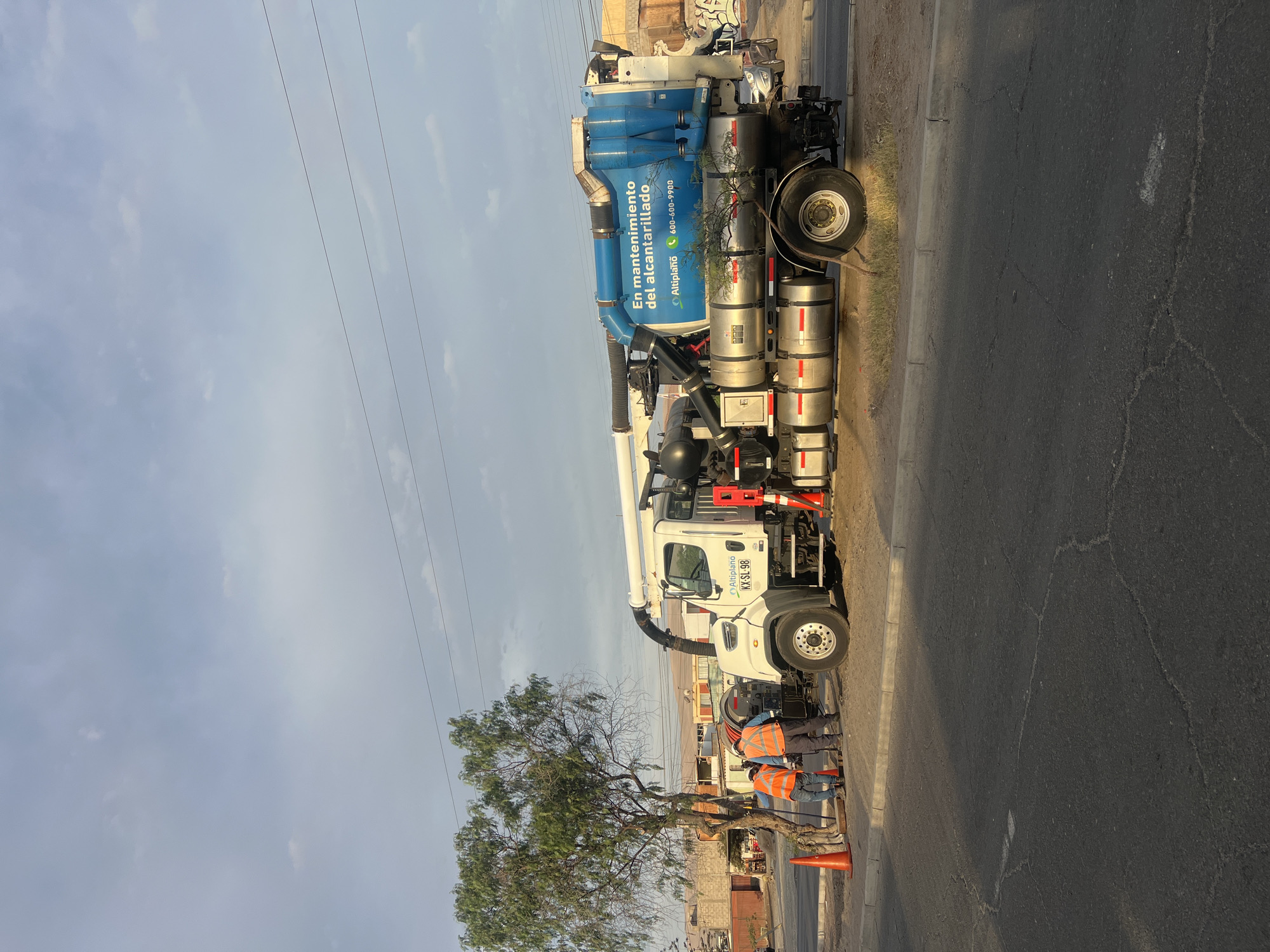 The image depicts a street scene in Arica, Chile, featuring a large truck used for sewer maintenance ("En mantenimiento del alcantarillado"). The truck, primarily white with a blue tank on the back, has the logo "Altiplano" and a phone number displayed on the tank. The truck is parked on the side of the street, with at least two workers wearing orange safety vests standing near the truck and what appears to be an open utility hole. An orange cone marks the area. A tree is situated nearby. In the background, there are buildings, with power lines stretching across the sky. The sky is overcast. The ground is cracked in multiple locations.