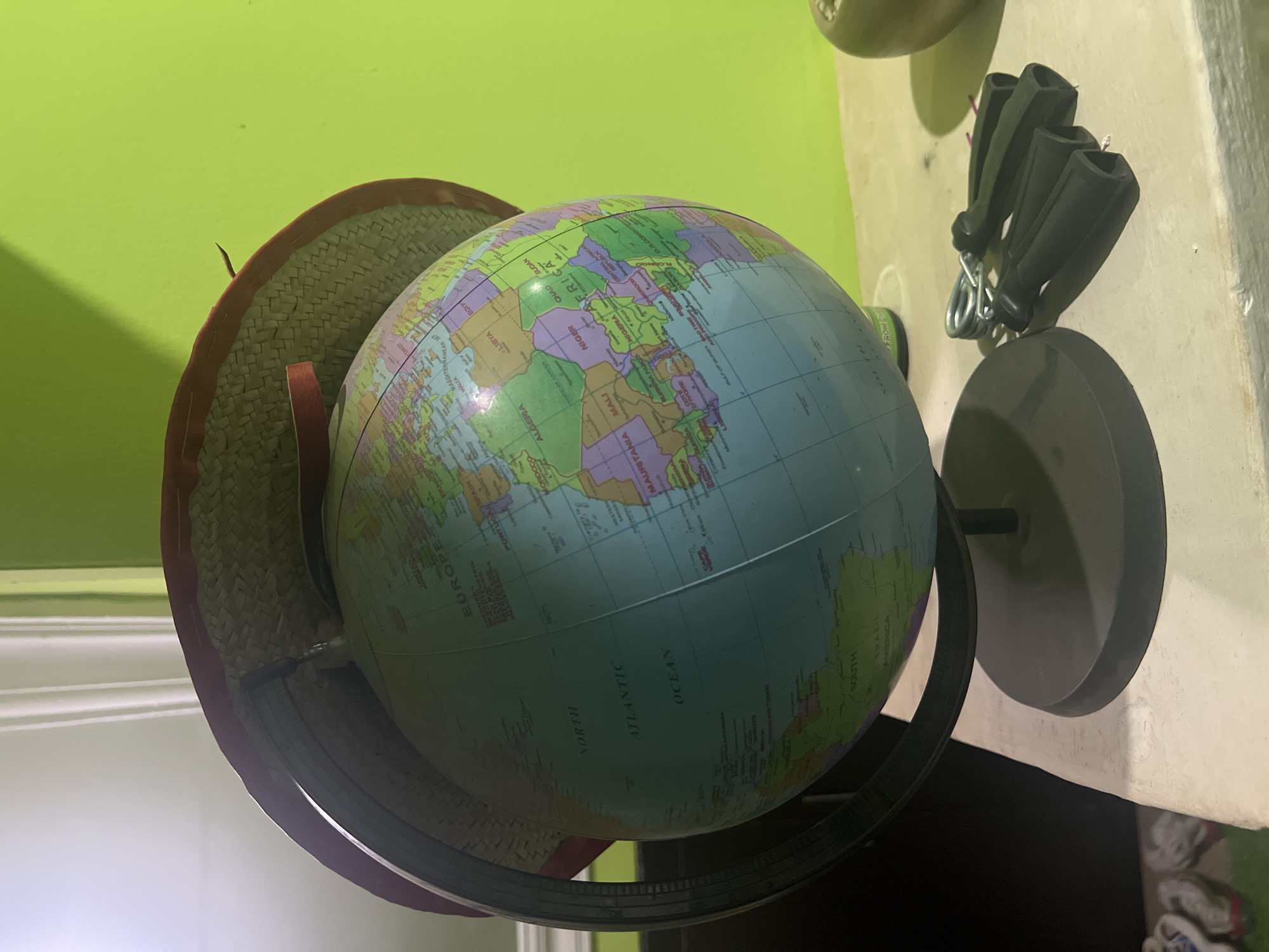 The image shows a globe resting on a stand. A straw hat is perched on top of the globe. The globe displays a map with labeled continents and oceans, including Africa and the Atlantic Ocean. The scene is lit by an unknown source. The location, Abuja, Nigeria, cannot be determined from the image.
