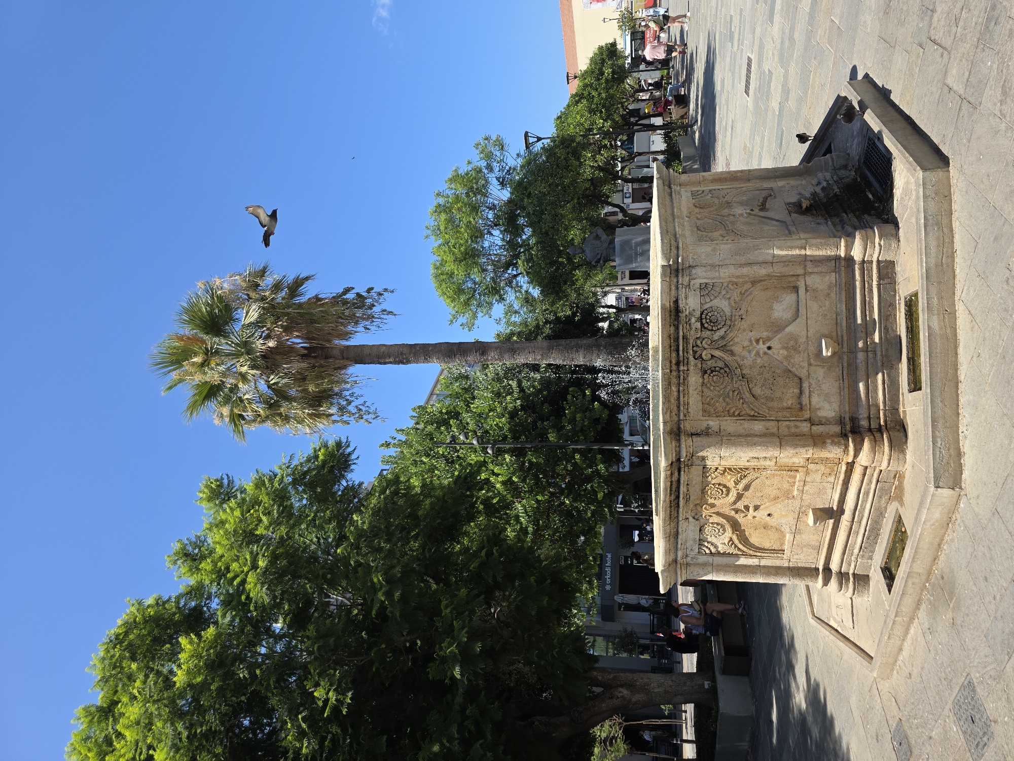 A pigeon flies in a bright blue sky above a stone fountain in the city of Chania, Greece. The fountain is ornate, with carved designs on its sides. A tall palm tree stands behind the fountain, and lush green trees line the background, with buildings and a street visible. Several people are seated on benches and walking in the distance. The ground is paved with stone tiles.