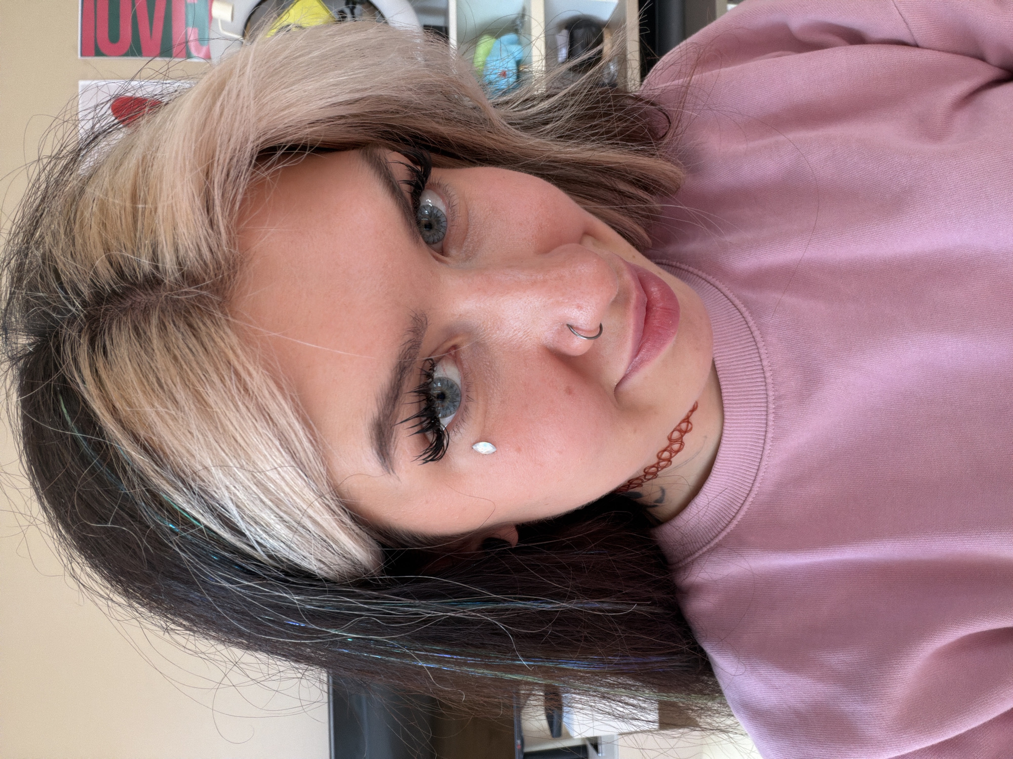 A close-up, rotated view of a woman with dyed hair (blonde and dark brown with sparkly strands) and blue eyes. She is wearing a light pink sweater. She has a nose ring and a possible decorative stone on her cheek. There is what appears to be a henna-like design on her neck. The background shows what could be shelves. The location in the city of Budva, Montenegro is not discernible from the image.