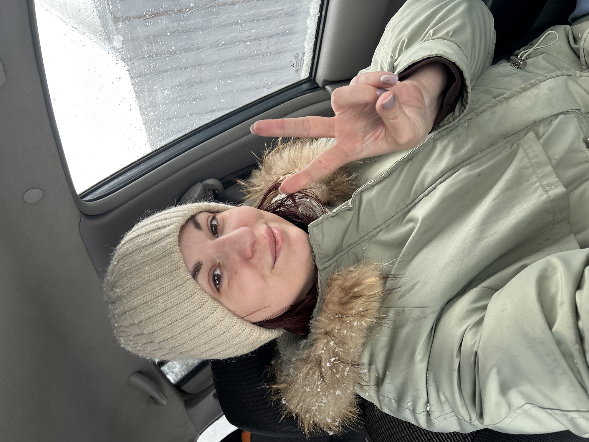 A woman is lying in a car, wearing a beige knit hat and a light green winter jacket with a fur-trimmed hood. She is holding up a peace sign with her right hand. Snow or rain is visible on the car's sunroof. The image was taken in the city of Фоки, Russia.