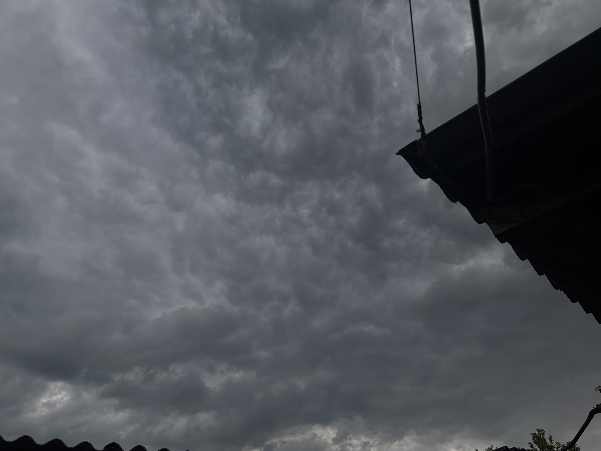 The image shows a stormy, overcast sky filled with dark, textured clouds. A portion of a dark, corrugated roofline is visible in the upper right corner, extending downwards and to the left, creating a strong diagonal line across the frame. A thin, dark rope hangs from a point above the roof, also extending downwards into the frame. In the lower portion of the image, another dark, wavy roofline can be seen, and a small glimpse of green foliage suggests trees or bushes beneath it. The overall scene evokes a sense of impending rain or a storm, with the dark clouds and muted lighting contributing to a somber or dramatic mood. The location is identified as Buky, Ukraine. No people are visible, and no text is present in the image. The time of day appears to be late afternoon or early evening, given the dark sky, but it is difficult to determine precisely due to the heavy cloud cover.