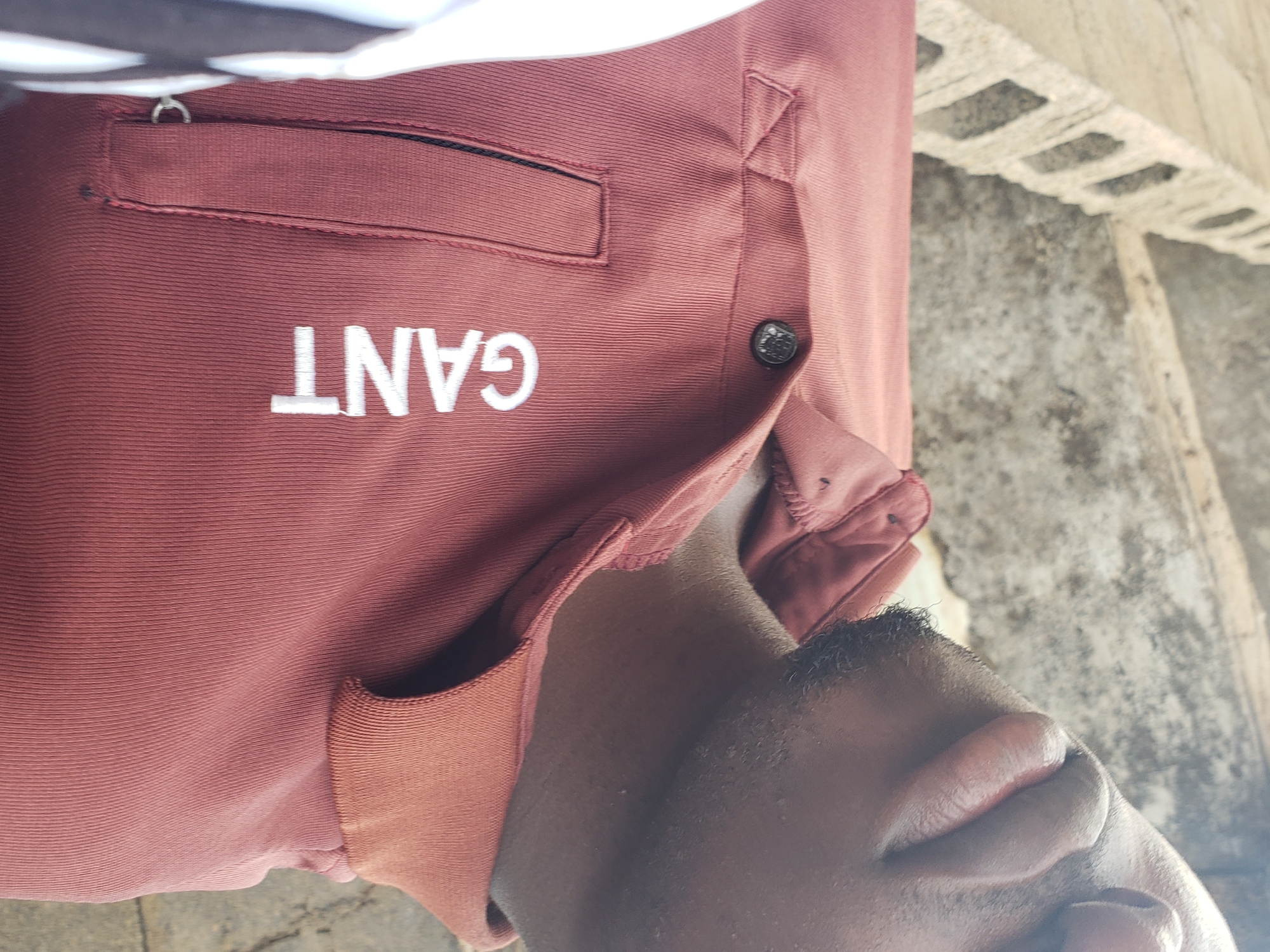 A close-up, chest-level, outdoor, eye-level shot focuses on the upper chest and chin area of a young Black man wearing a dark red polo shirt. The shirt has a ribbed collar and a pocket with a dark trim. White lettering on the shirt reads "GANT". The man has a slight beard and is looking slightly down and to the left. Behind him, a rough, concrete block wall runs horizontally across the frame, with a textured, gray concrete wall in the background. The lighting suggests it is daytime. The scene appears to be in the city of Kachia, Nigeria.