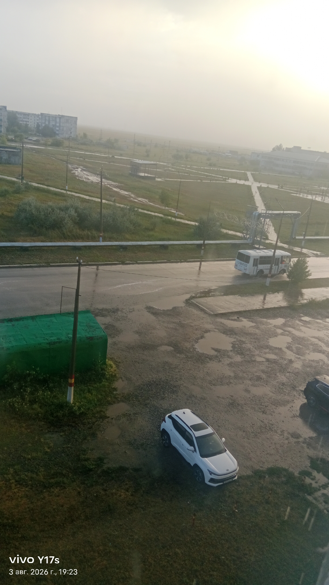 The image shows a street scene in Rudny, Kazakhstan, on a hazy or foggy day, possibly during or after rain. In the foreground, a white SUV is parked on a muddy surface next to a grassy area. Further down the street, a white bus is in motion, splashing water. A green structure is visible on the left side of the road. In the background, apartment buildings and a sports facility with a partially constructed roof are discernible through the atmospheric haze. The overall impression is one of overcast weather and a somewhat underdeveloped urban environment.