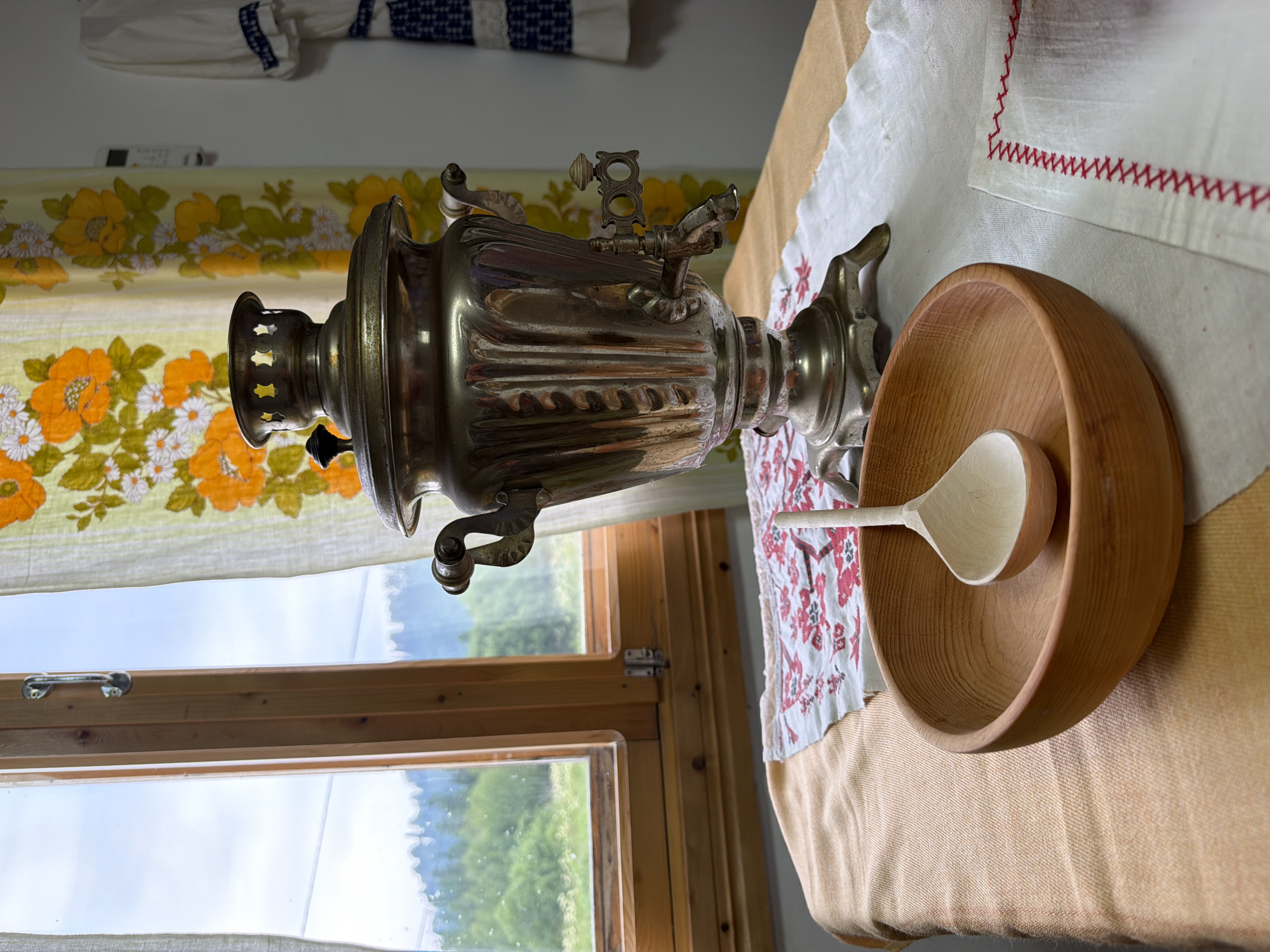 A close-up, eye-level shot captures a still life arrangement on a table with a window and floral curtains in the background. The table is covered with an orange cloth, a white cloth with red embroidery, and a piece of patterned fabric. In the foreground, a wooden bowl contains a wooden ladle. Behind the bowl, a silver samovar stands on a decorative base, featuring a spigot and handles. The window behind the samovar reveals a verdant, hilly landscape with trees. The curtains, patterned with orange and yellow flowers, hang to the right of the window. On the wall to the far right, a piece of embroidered clothing is visible. The scene suggests an indoor setting in Ust'-Kabyrza, Russia, possibly a home or a traditional craft shop, with elements of domesticity and regional culture.