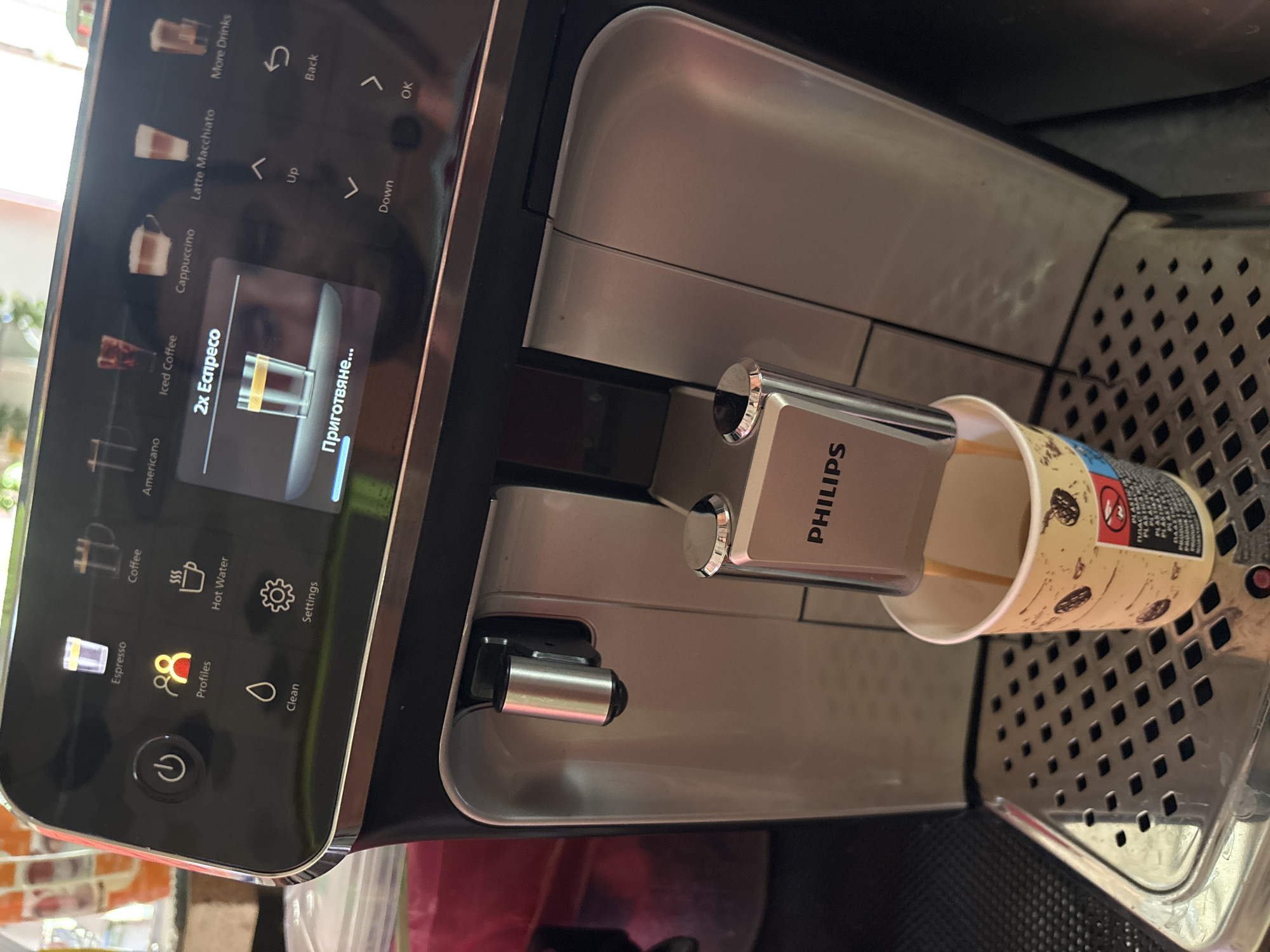 The image shows a Philips coffee machine making a double espresso into a paper cup. The machine's display indicates the process is underway ("Приготвяне..." which means "Preparing..." in Bulgarian), and the selection is for "2x Еспресо" (2x Espresso). The user interface includes other drink options like Cappuccino and Latte Macchiato. The setting appears to be in a domestic environment. While not discernible from the image, it is mentioned to be in Gorna Lipnitsa, Bulgaria.