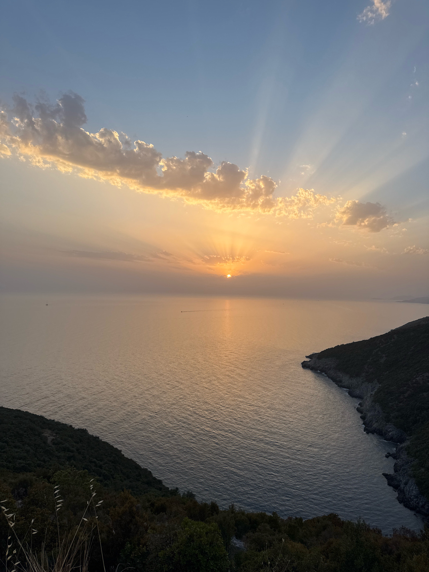 A high-angle, wide shot captures a serene sunset over the Adriatic Sea, likely near the city of Zagora, Montenegro. The sun, partially obscured by wispy, golden-edged clouds, casts dramatic crepuscular rays across the pale orange sky. The sea stretches out, reflecting the soft light, with subtle ripples indicating a gentle breeze. In the distance, two small boats, one appearing to be a sailboat and the other a motorboat, are visible as dark specks on the water. The foreground is dominated by lush, dark green vegetation covering the sloping hillsides that frame the bay. The rugged coastline, with its rocky outcrops, meets the water's edge. The overall scene evokes a sense of tranquility and natural beauty.