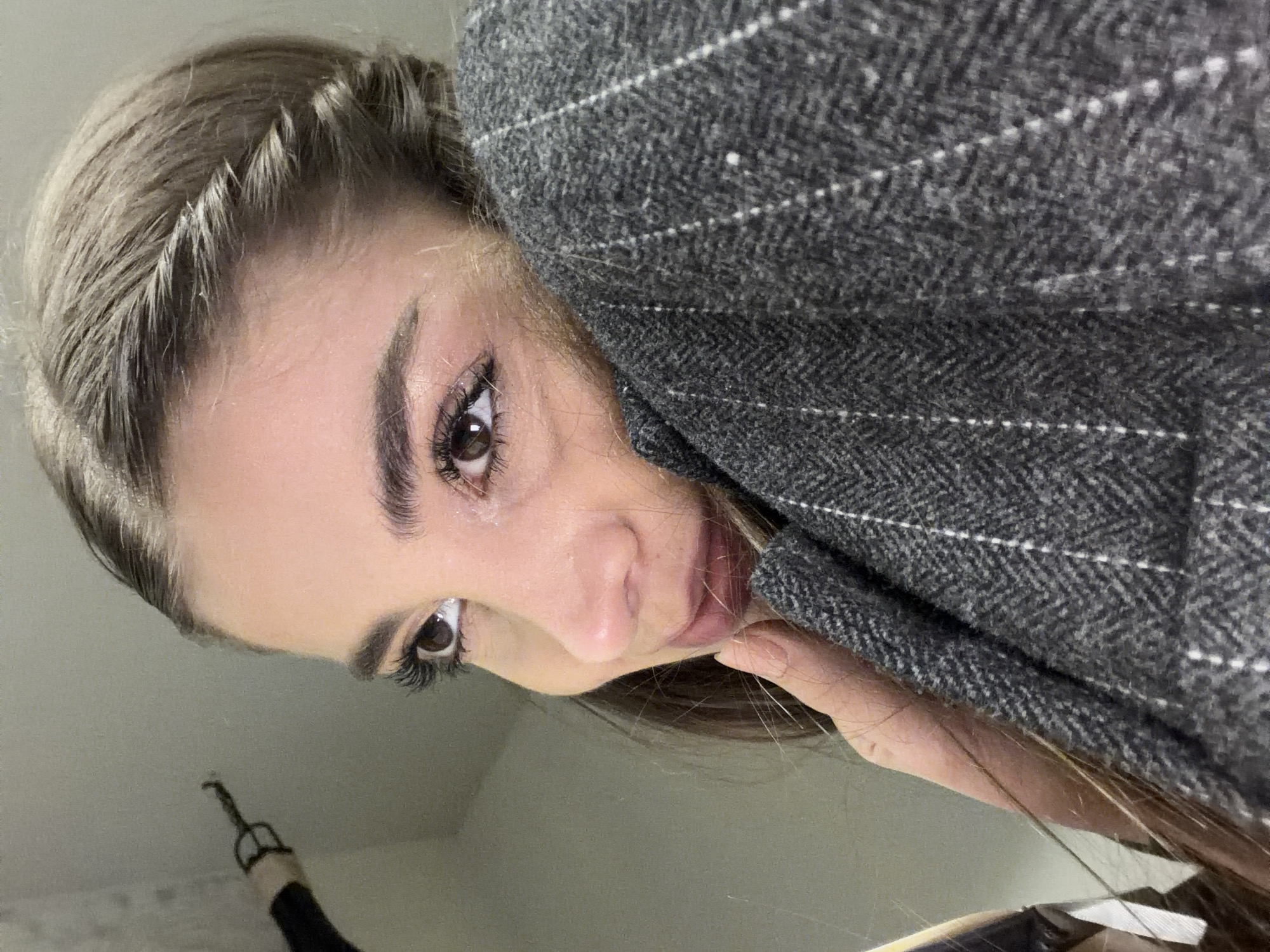 A close-up, tilted portrait shows a young woman looking directly at the camera. Her head is angled, giving the impression of an upward gaze or an inverted perspective. She has light brown hair, styled with a small, visible braid near her forehead on the left side of her face. Her large, dark brown eyes are framed by well-defined eyebrows and prominent eyelashes. Her lips are a light pink, slightly parted, with her fingers gently resting near her chin.
She is wearing a thick, textured gray jacket or coat, possibly tweed or wool, featuring a herringbone weave with subtle white pinstripes. The background is a plain, light-colored surface, likely a wall or ceiling. In the lower left corner of the image, a dark object resembling a bag handle with a metallic clasp is partially visible. The photograph appears to be a casual selfie. There is no visible text. The location context for this media file is Yerevan, Armenia.