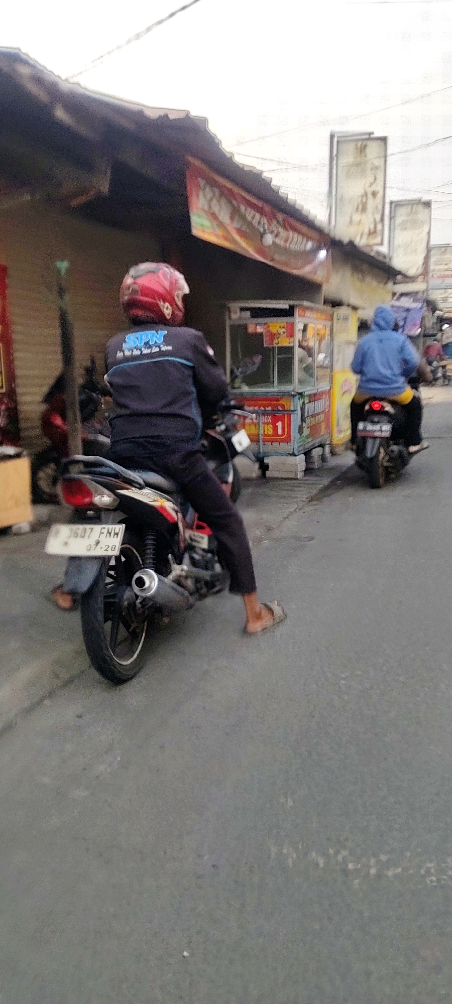 In Mekarmukti, Indonesia, a medium-shot outdoor photograph captures two individuals on motorcycles on a paved road. The foreground features a person wearing a red helmet and a dark jacket with "SPN" in blue lettering, seated on a black motorcycle. The motorcycle's license plate is visible, displaying "B 3607 FNW" and "07-28". The rider's legs are visible, wearing dark pants and open-toed sandals. To the right, another person wearing a blue hooded jacket and yellow pants rides a black motorcycle. This motorcycle has a red license plate. Behind the motorcycles, a food cart with a glass display case is positioned. The cart is adorned with colorful signage, including the text "Makan" and "Gratis 1". In the background, a building with a corrugated metal roof and awnings is visible, along with a few other structures and people. The sky is overcast.