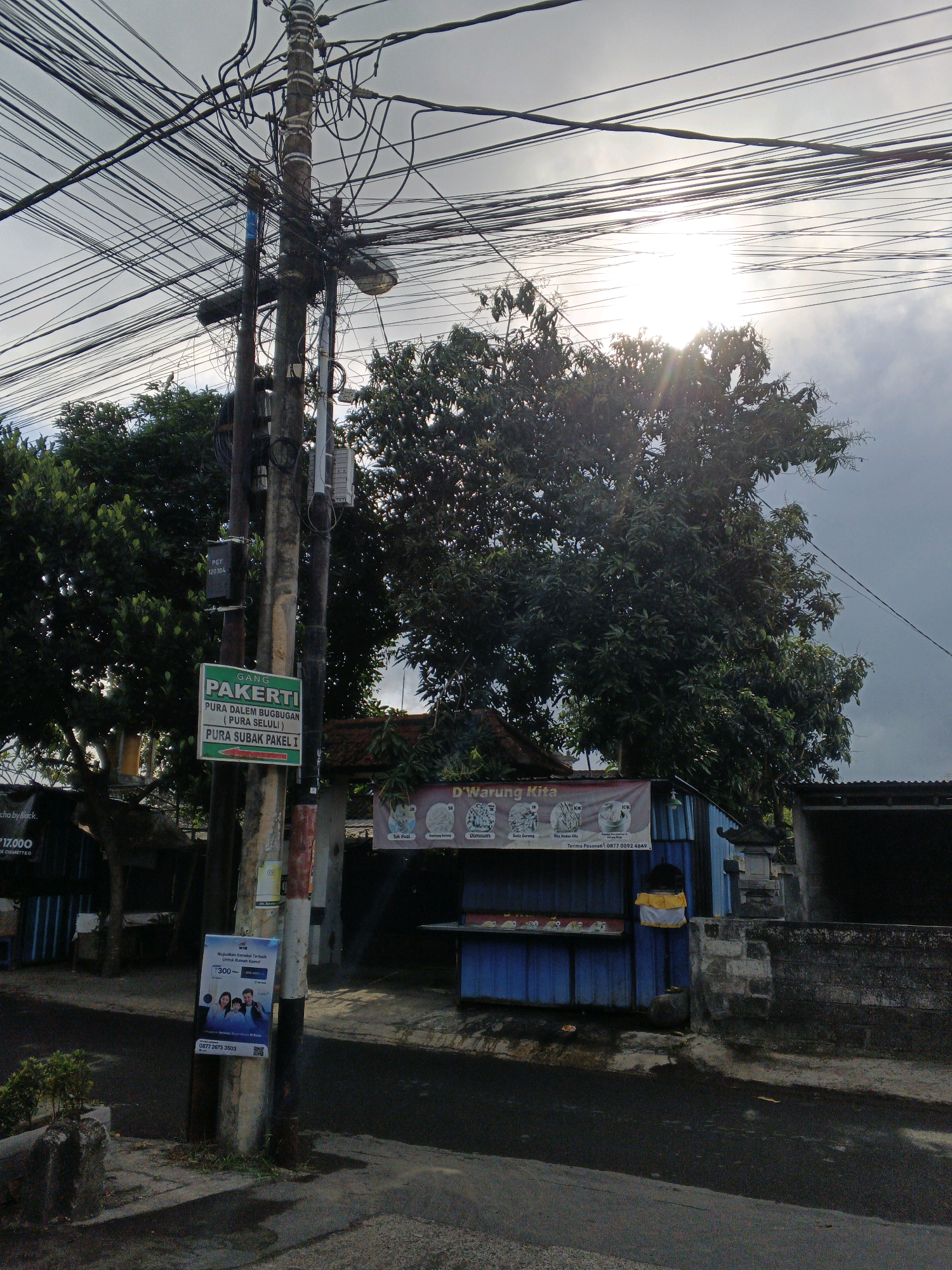 The image captures a street scene in Denpasar, Indonesia, likely on a cloudy day with sunlight filtering through the overcast sky. A prominent utility pole, heavily laden with a dense network of electrical wires, stands in the foreground. Attached to this pole are several signs, including a green sign with white text that reads "GANG PAKFERTI" and further instructions in Indonesian pointing to "PURA DALEM BUGBUGAN (PURA SELULI)" and "PURA SUBAK PAKEL I." Below this, a blue and white poster depicts a family and text related to a financial product or service. Further down the pole, a small, dark rectangular box with the label "PGY 120304" is affixed.
To the right of the utility pole, a blue building serves as a small food stall named "WARUNG KITA." A banner above the stall displays various food items with prices in Indonesian Rupiah. The stall has a blue corrugated metal facade with a counter displaying food behind a clear barrier. A yellow and white cloth is draped over the side of the stall. Behind the stall, a stone wall and more trees are visible. The street in front of the stall appears to be paved and slightly wet. The overall environment suggests a typical urban streetscape in Bali, characterized by dense electrical infrastructure and roadside eateries.
