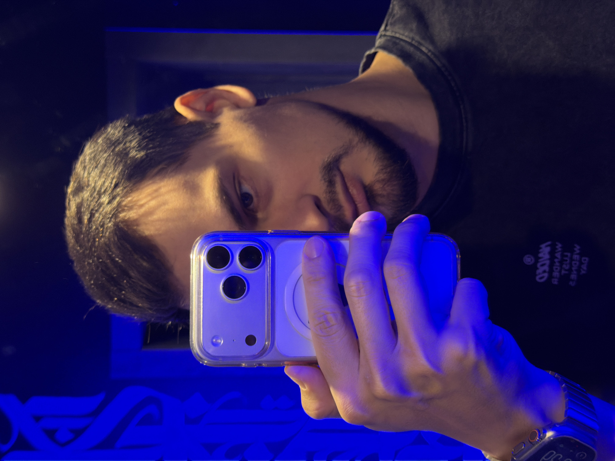 A man is taking a selfie in a mirror with his iPhone. The man has dark hair and a beard. He is wearing a black shirt and an Apple watch. The environment has blue lighting. The location is unknown and there is no indication of it being Krasnoyarsk, Russia.