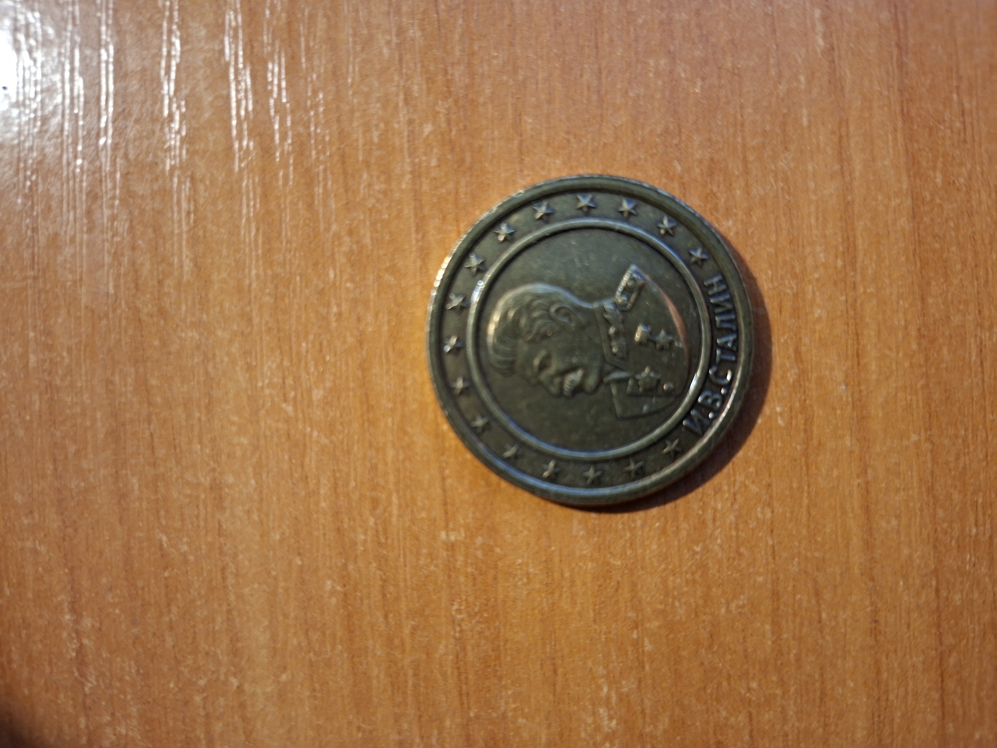 A metallic coin with a relief of Joseph Stalin is displayed on a wooden surface. The coin is circular and has a raised border of small stars. Stalin's profile is depicted facing right, wearing military attire. Below the profile, Russian text is inscribed. The wooden surface has a visible grain pattern and appears to be a table or desk. The lighting suggests an indoor environment, possibly with overhead light casting reflections on the polished wood.