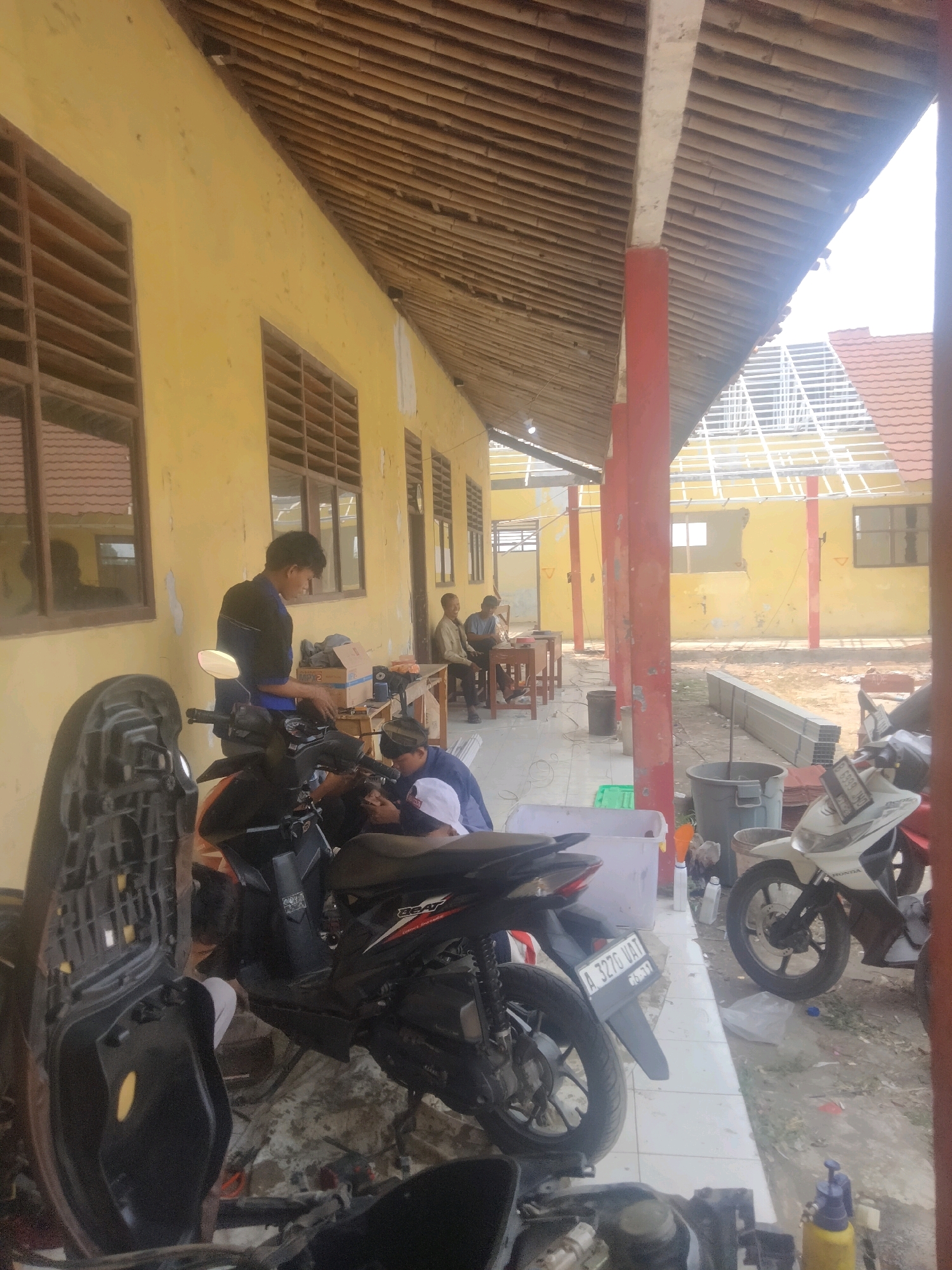 The image depicts an outdoor scene in front of a yellow building with a thatched roof. Several individuals are present, some appearing to be working on motorcycles. In the foreground, two motorcycles are partially visible, with one in the process of being dismantled or repaired. A person in a blue shirt is leaning over one of the motorcycles. Further back, a young man in a dark shirt is standing near a table with a cardboard box. Two other men are seated at a table in the background. The environment appears to be somewhat unkempt, with building materials and debris scattered around. The overall impression is one of a repair or construction area associated with the building.
