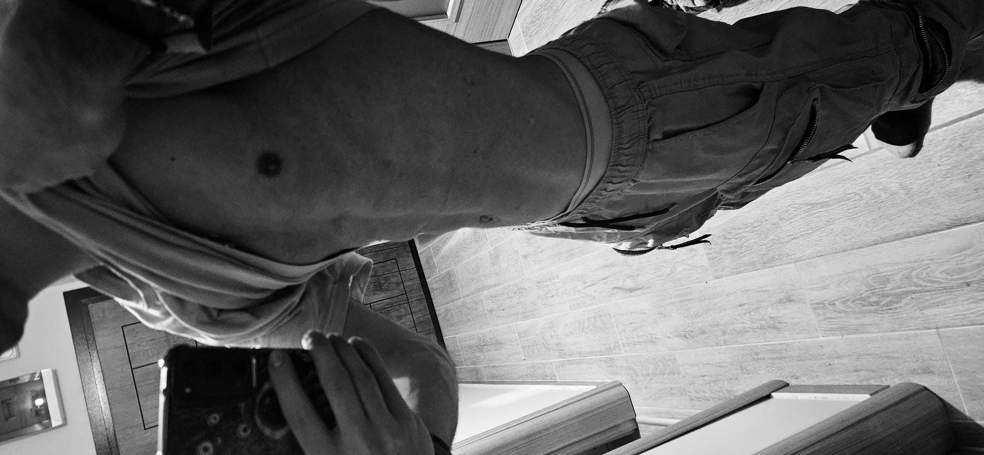 A shirtless person wearing pants and holding a phone is taking a mirror selfie. The person's torso is visible, and they are wearing a t-shirt. The background shows a tiled floor, a door, and furniture, possibly including a mirror. The location is unknown, and there is no indication that it is in Plovdiv, Bulgaria. There are no identifiable interactions beyond the selfie.