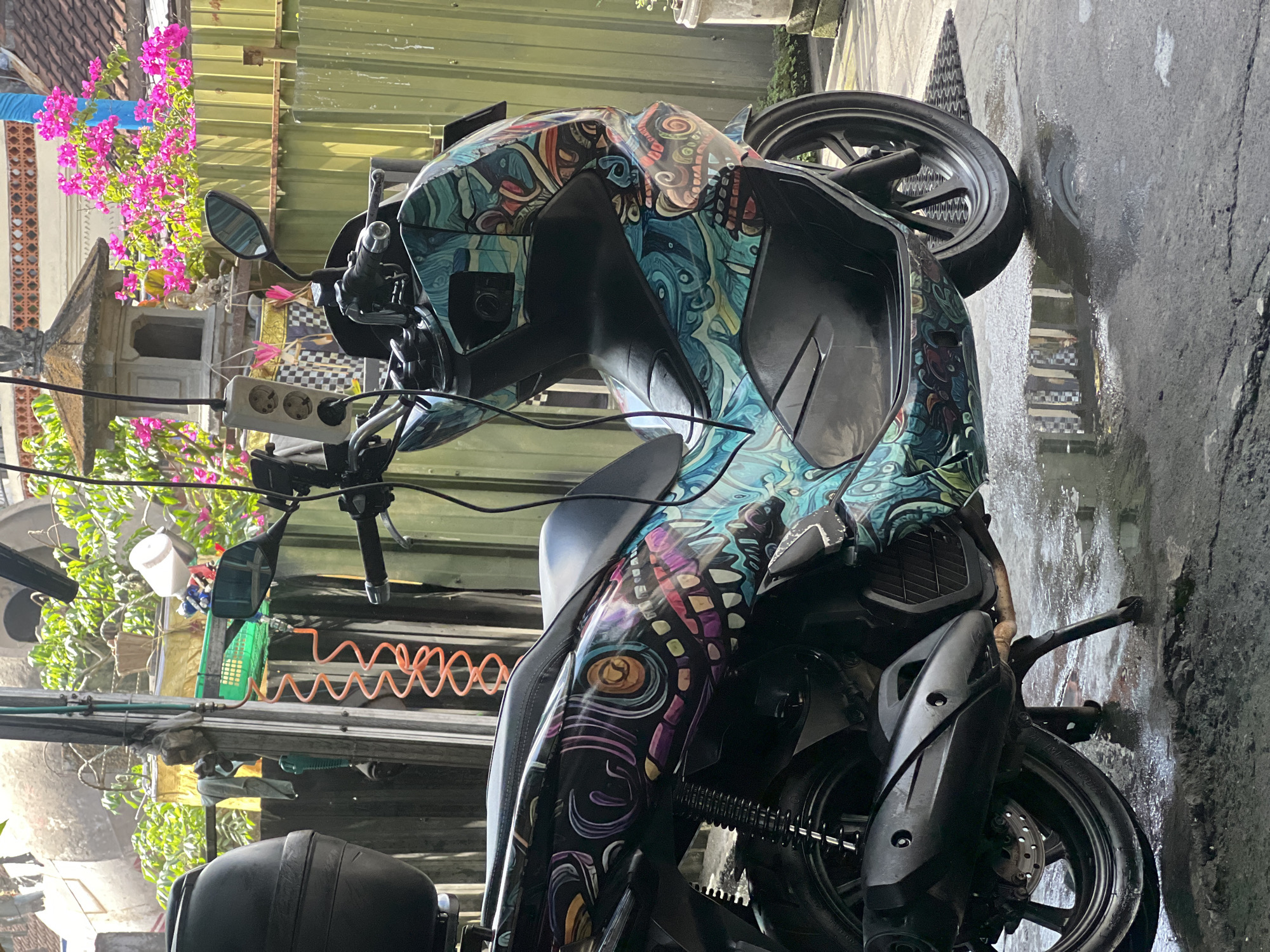 A scooter with a custom, colorful, psychedelic design is parked on a wet concrete surface in what appears to be a residential or workshop area. The scooter has a black seat, black storage box on the back, and a custom exhaust pipe. The design features swirling patterns, eye-like motifs, and a skull-like image in shades of blue, teal, pink, purple, and orange. In the background, a green corrugated metal fence is visible, with a small shrine or decorative structure and pink bougainvillea flowers above it. There are also some wires and a coiled orange hose hanging from a pole. The ground is wet and reflects some of the surroundings, suggesting it may have recently rained or been washed. There are no people visible in the image.