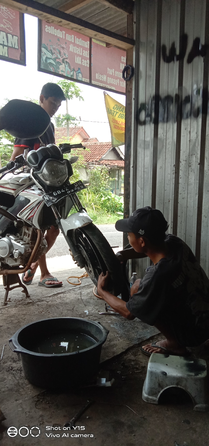 This image captures a motorcycle repair scene within an open-fronted shop in Ranugedang, Indonesia. Two adult males are present.
One male, wearing a dark t-shirt and a black baseball cap, is kneeling barefoot on a stained concrete floor, actively working on the front tire of a white and black Yamaha motorcycle. He is manipulating the tire and rim with his hands, suggesting a repair or replacement. A large black plastic basin filled with clear liquid, along with various metal tools, rests on the floor near him. A small white plastic stool is also visible on the ground.
The second male, dressed in a dark t-shirt with red accents and wearing light-colored flip-flops, stands partially obscured behind the motorcycle's handlebars. He appears to be observing the repair. The motorcycle is on its kickstand, and its front license plate, "QR 6842 03-22," is clearly visible.
The shop interior features a corrugated metal wall on the right, bearing dark, indistinct graffiti-like text. Overhead, wooden beams and a corrugated metal roof structure are present. Several inverted red signs with white Indonesian text advertising services such as "Tambal Ban Tubeless," "Las Listrik," "Gas Aki," and "Ganti Oli" are hanging within the shop's opening. A partially visible yellow banner with red text is also seen outside. Beyond the shop, a paved surface leads to residential buildings with orange-red tiled roofs, surrounded by dense green foliage, under a bright, possibly overcast, sky.