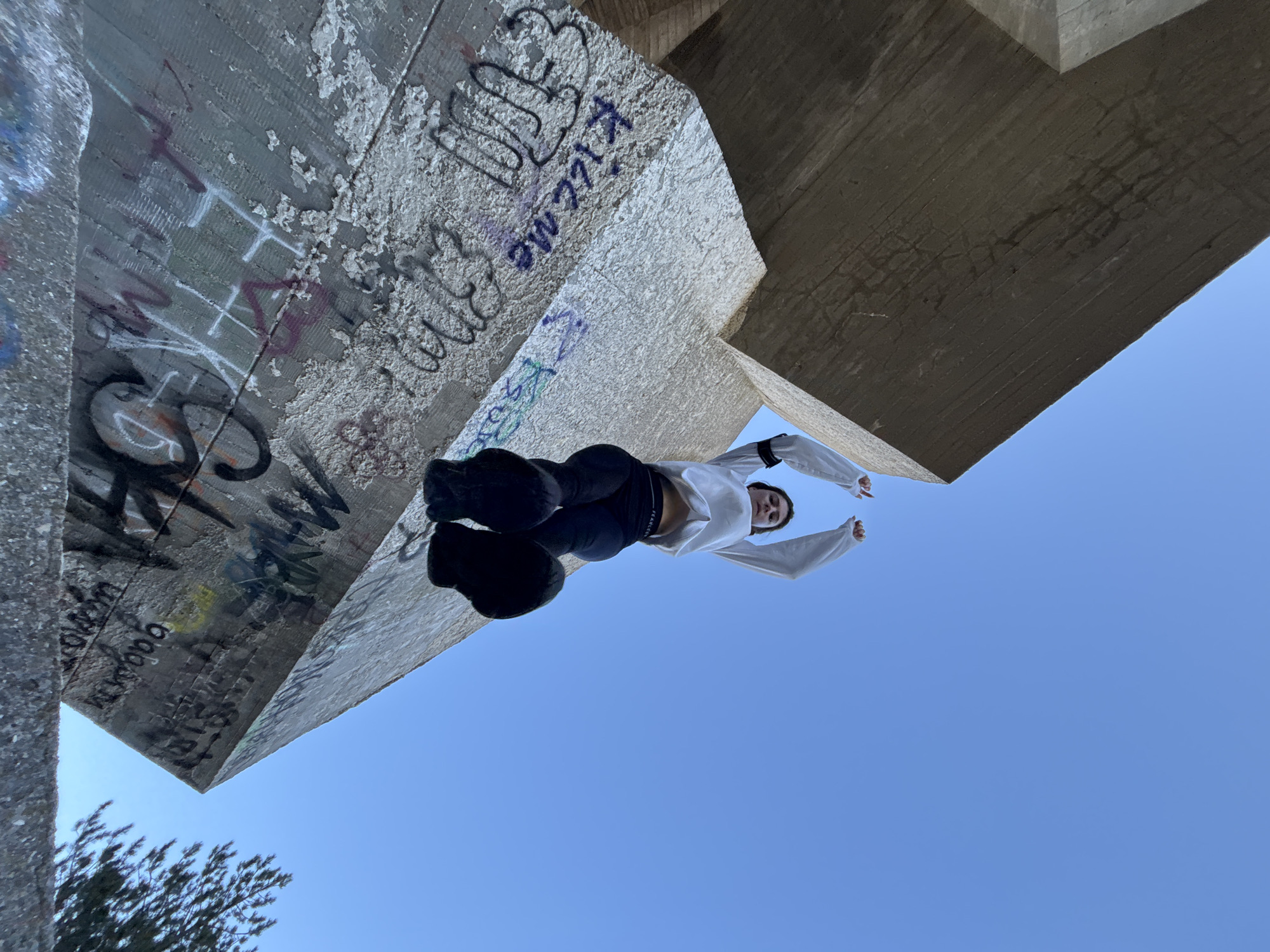 A low-angle shot looking up at a person standing atop a concrete structure. The person is wearing a white long-sleeved shirt and dark pants. The sky is clear and blue, filling the majority of the frame. The concrete structure features graffiti. The location is in Varna, Bulgaria.