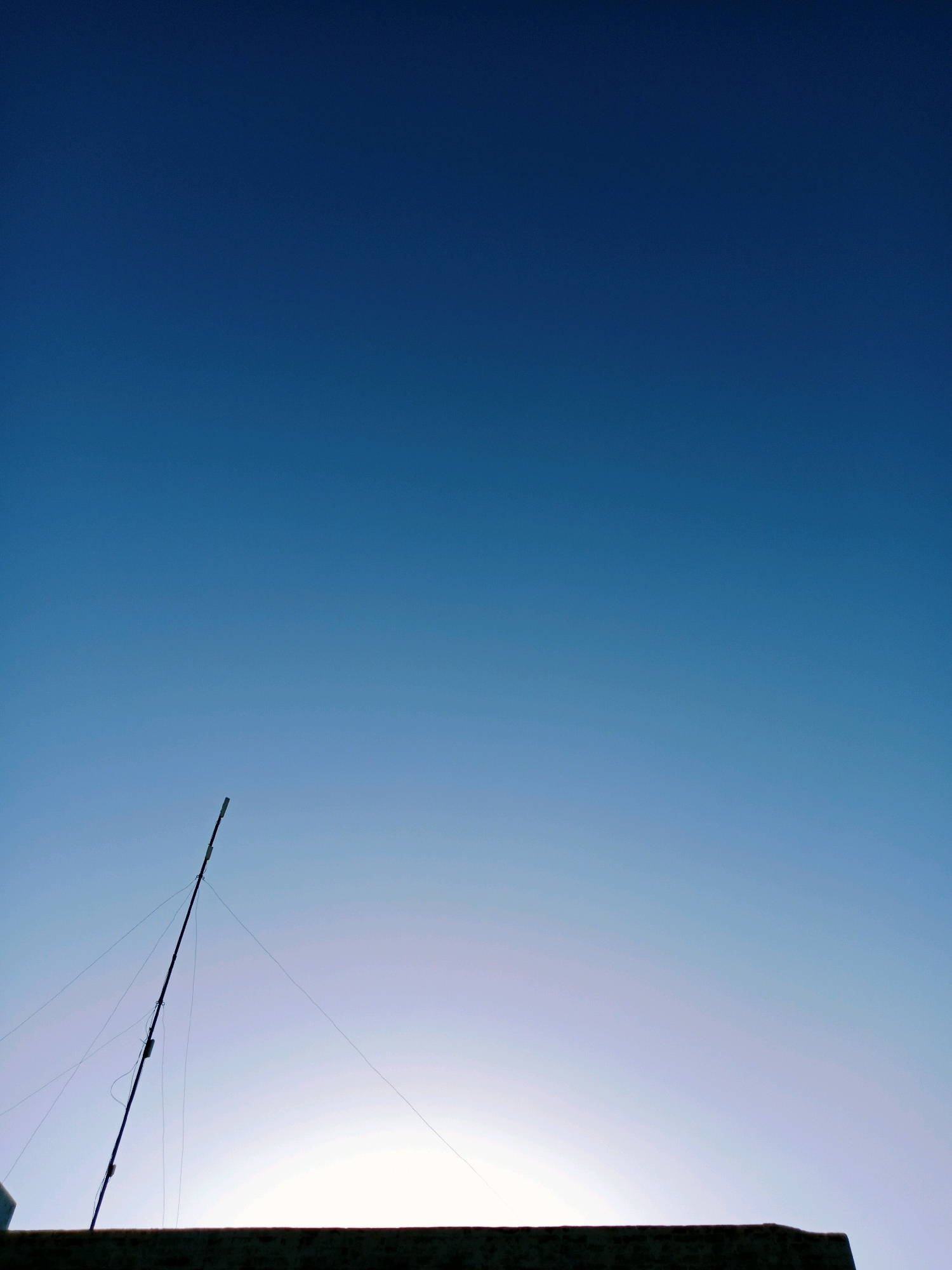 A tall, thin antenna pole leans against the bright sky, its surface punctuated by several small, dark objects. Wires extend from the pole, fanning out and disappearing towards the right and left. Below the sky and the antenna, a dark, textured surface, likely the top of a building or wall, stretches across the bottom of the frame. The sky is a gradient of deep blue at the top, fading to a lighter blue and then a pale, hazy white at the bottom, suggesting either dawn or dusk. The overall impression is one of urban infrastructure against a vast, atmospheric sky. The location is Bahawalpur City Tehsil, Pakistan.