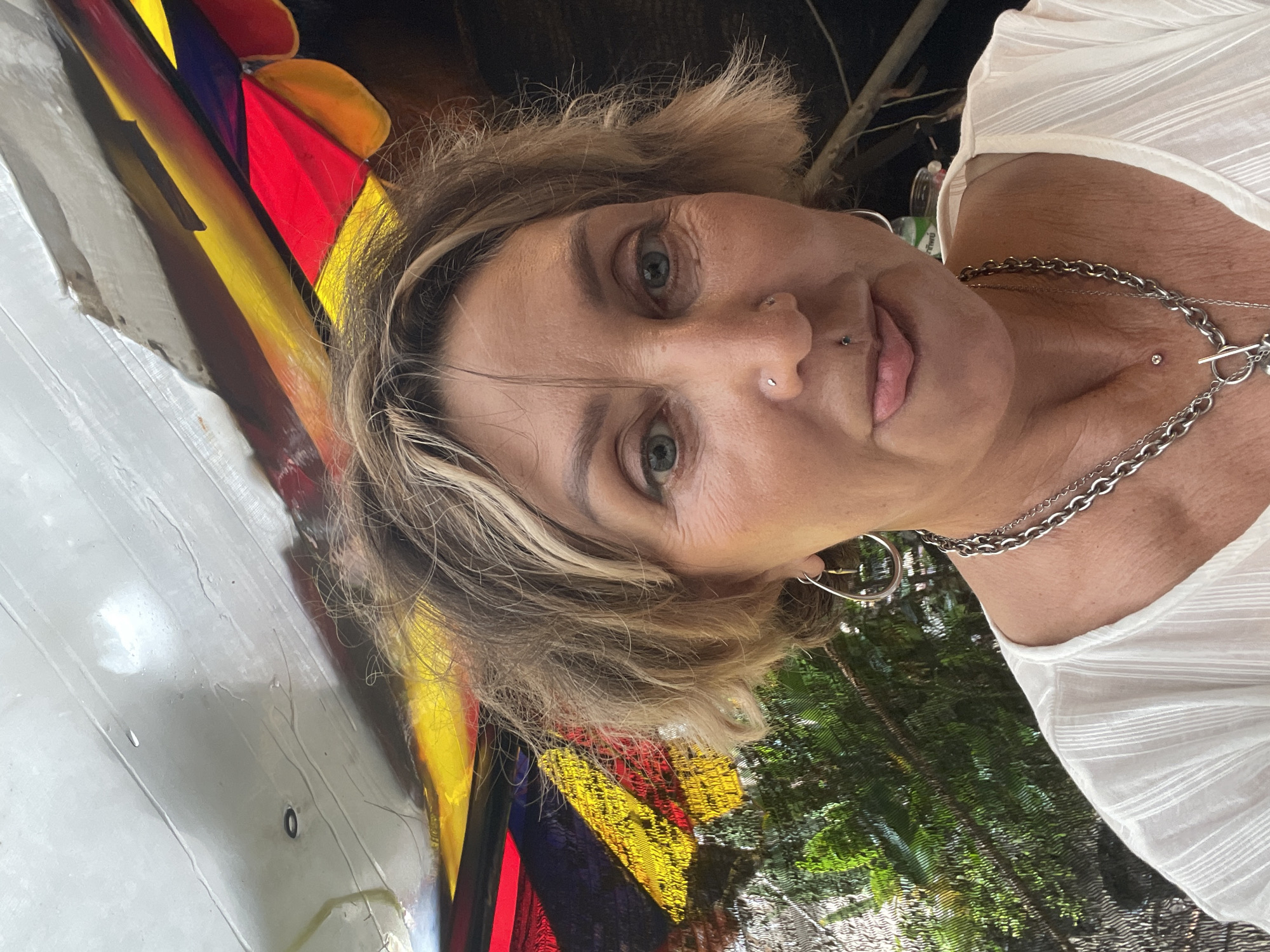 A woman with blonde, short, messy hair, blue eyes, and light skin is taking a selfie outdoors. She has piercings in her nose and lip, and is wearing large hoop earrings and a silver chain necklace. She is wearing a white top with textured fabric. Behind her, a colorful awning made of red, yellow, and blue fabric is visible, along with greenery. The setting appears to be a casual outdoor area, possibly a cafe or market, in Choeng Thale, Thailand.