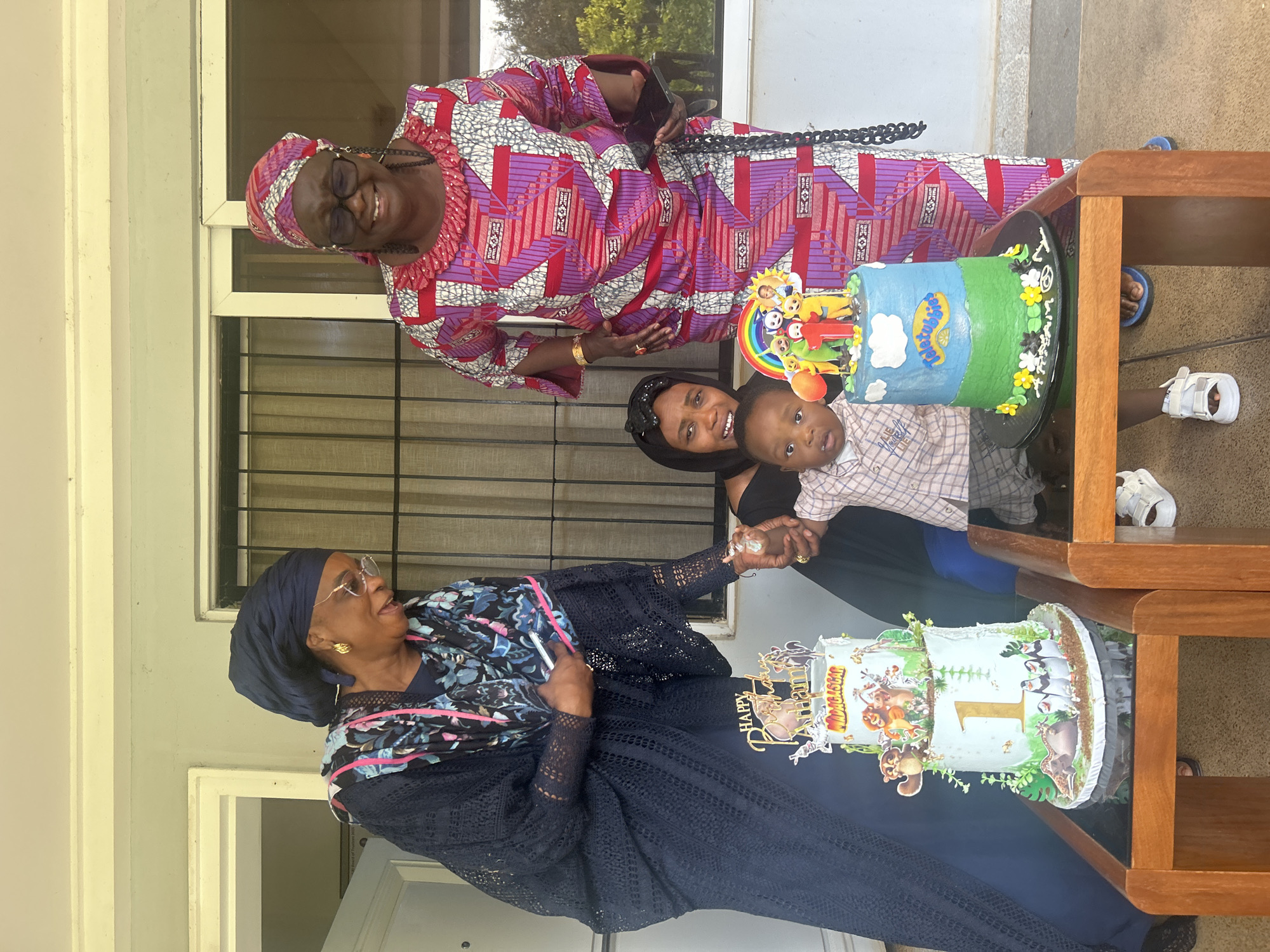 In Dutsen Kato, Nigeria, a two-tiered birthday cake with Madagascar characters stands on a glass-topped wooden table. To the left, a woman in a dark blue dress and headscarf leans over the cake, holding a phone. To her right, another woman in a vibrant pink and purple patterned dress and headscarf smiles. In front of the cakes, a woman in a black headscarf holds a baby wearing a plaid shirt and white sandals. The background features a building with windows and vertical bars.
