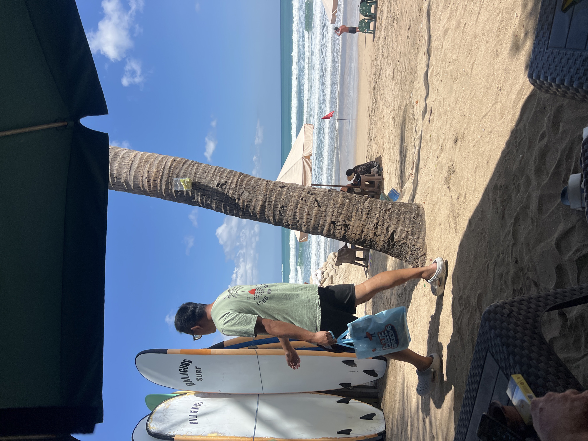 The image captures a sunny day at Kuta Beach, Bali, Indonesia, with a focus on a man walking on the sand. He is wearing a green t-shirt, black shorts, and grey sandals, carrying a light blue tote bag with the words "BLUE GROOVE" printed on it. To his left, a row of white surfboards stands upright, some with branding like "BALI GUNS" and "SURF".
The background reveals the ocean with gentle waves and a clear blue sky with scattered white clouds. Palm trees are visible, with one prominently in the foreground casting a shadow. Several beach umbrellas and chairs are set up on the sand. In the distance, a red flag is planted in the sand near the water. A few other people are visible in the background, some seated under umbrellas. In the immediate foreground, a portion of a dark rattan table is visible, with a pack of cigarettes on it. The overall scene depicts a typical beach day at a popular Indonesian surf spot.