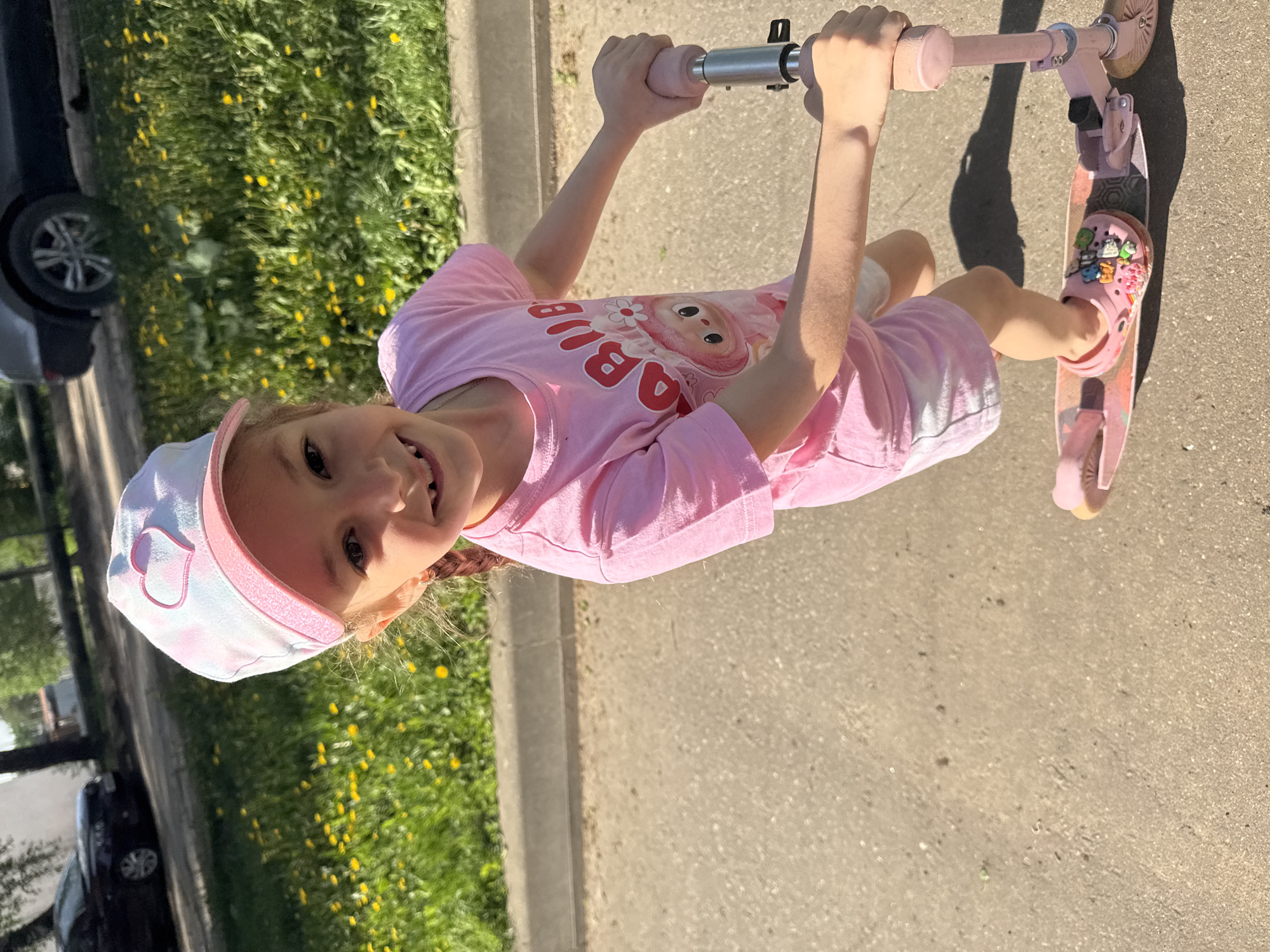 A young girl wearing a pink t-shirt and a pink and white cap is riding a pink scooter on an asphalt surface. She is smiling and looking at the camera. The location is outdoors and appears to be in the city of Раздолье, Russia, as evidenced by the street and vegetation along the curb. A car is parked alongside the curb.
