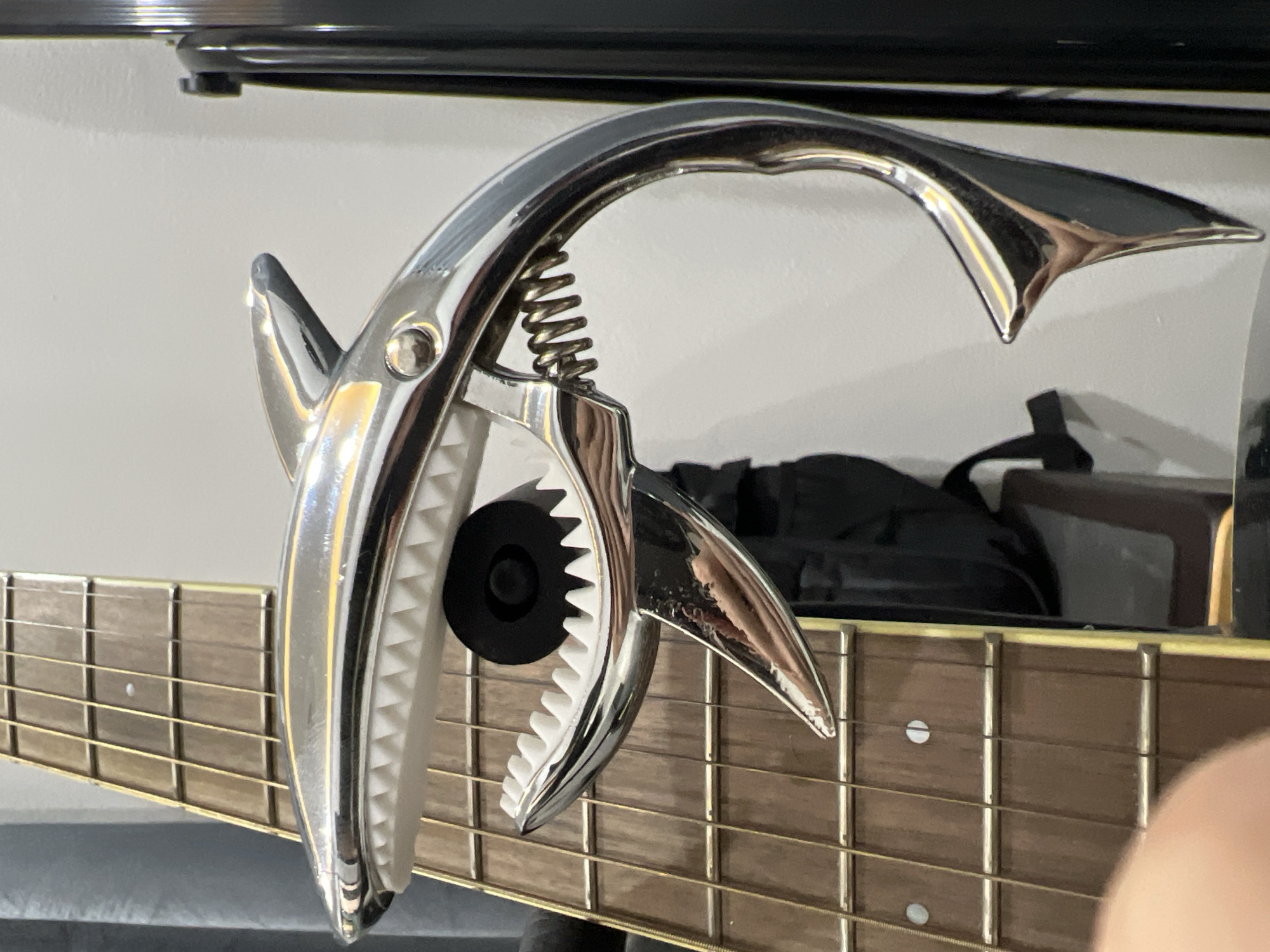 A chrome-plated guitar capo, shaped like a shark with its mouth open to reveal white teeth, is attached to the fretboard of an acoustic guitar. The capo is positioned over the strings on the guitar's neck. The guitar has a brown wooden fretboard with six strings and dot inlays. The background consists of a white wall and a dark vertical object, possibly a guitar stand or amplifier. The scene is indoors, and the lighting is neutral, casting soft reflections on the shiny surface of the capo. The location is not specified in the image, and therefore, cannot be determined to be in Choeng Thale, Thailand.
