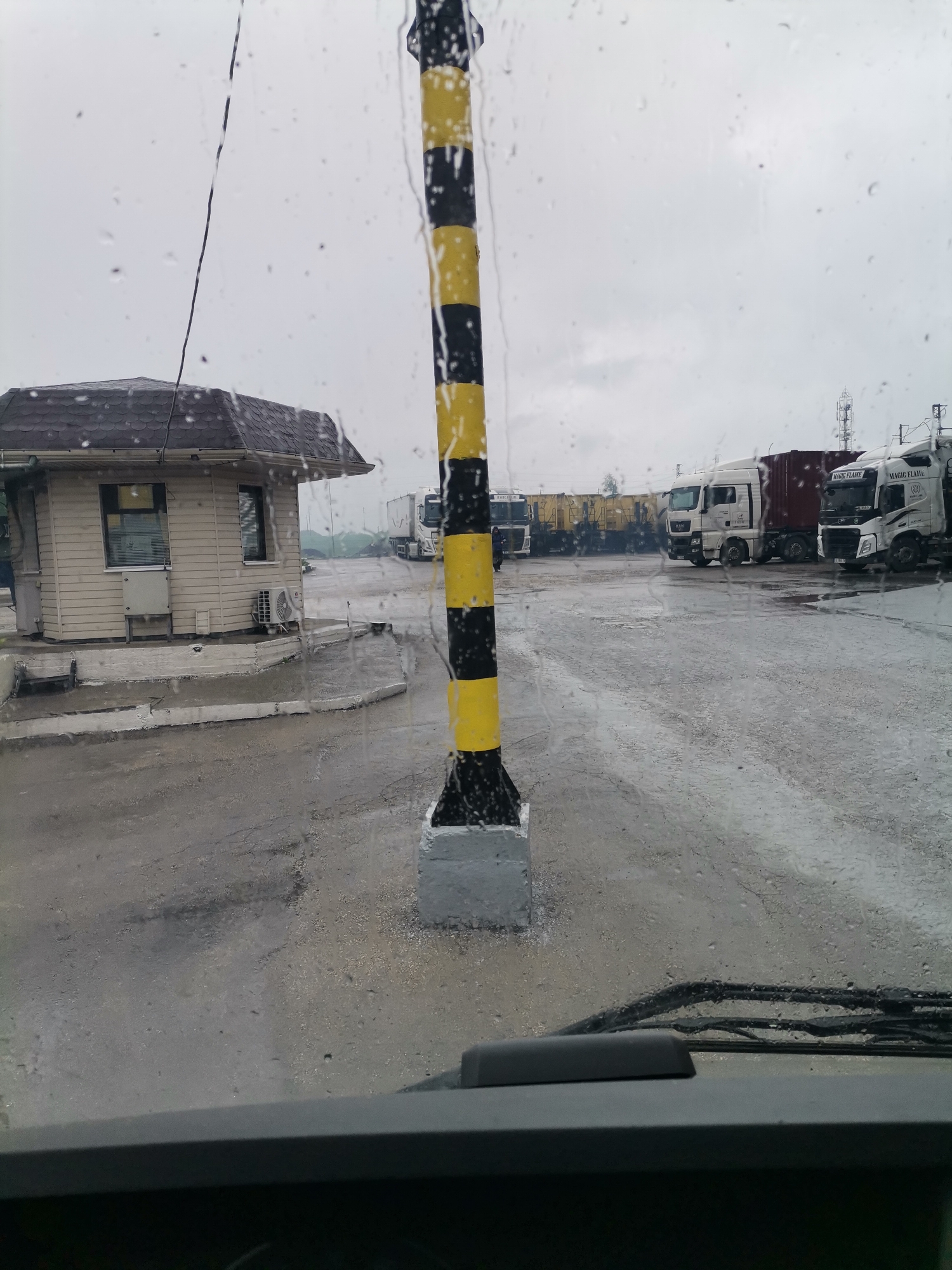 This image captures a damp, overcast day scene, viewed through a rain-streaked windshield from inside a vehicle. The foreground shows the top of a vehicle's dashboard and windshield wipers, with numerous raindrops distorting the view. A prominent black and yellow striped barrier post, anchored in a concrete base, stands vertically on the right side of the frame.
To the left, a small, light-colored guard house or security booth with a dark, shingled roof is visible. It features several windows, one with blinds, and an external air conditioning unit. The ground is a wet, paved surface, appearing slick from recent rain.
In the midground and background, an array of semi-trailer trucks are parked. Most of these are white cab-over trucks, some coupled with red trailers. Visible text on the trucks includes "MAN" and "MAGIC FLAME". Further in the background, railroad cars or wagons can be faintly seen. The sky is uniformly gray and cloudy, indicative of a dreary day. A faint, indistinct person is visible near a truck on the left.
The setting is an industrial or commercial yard, likely a logistics or transport center, characterized by the presence of trucks, a guard house, and a barrier. The wet conditions and muted lighting suggest it is daytime. The location context is Karapelit, Bulgaria.