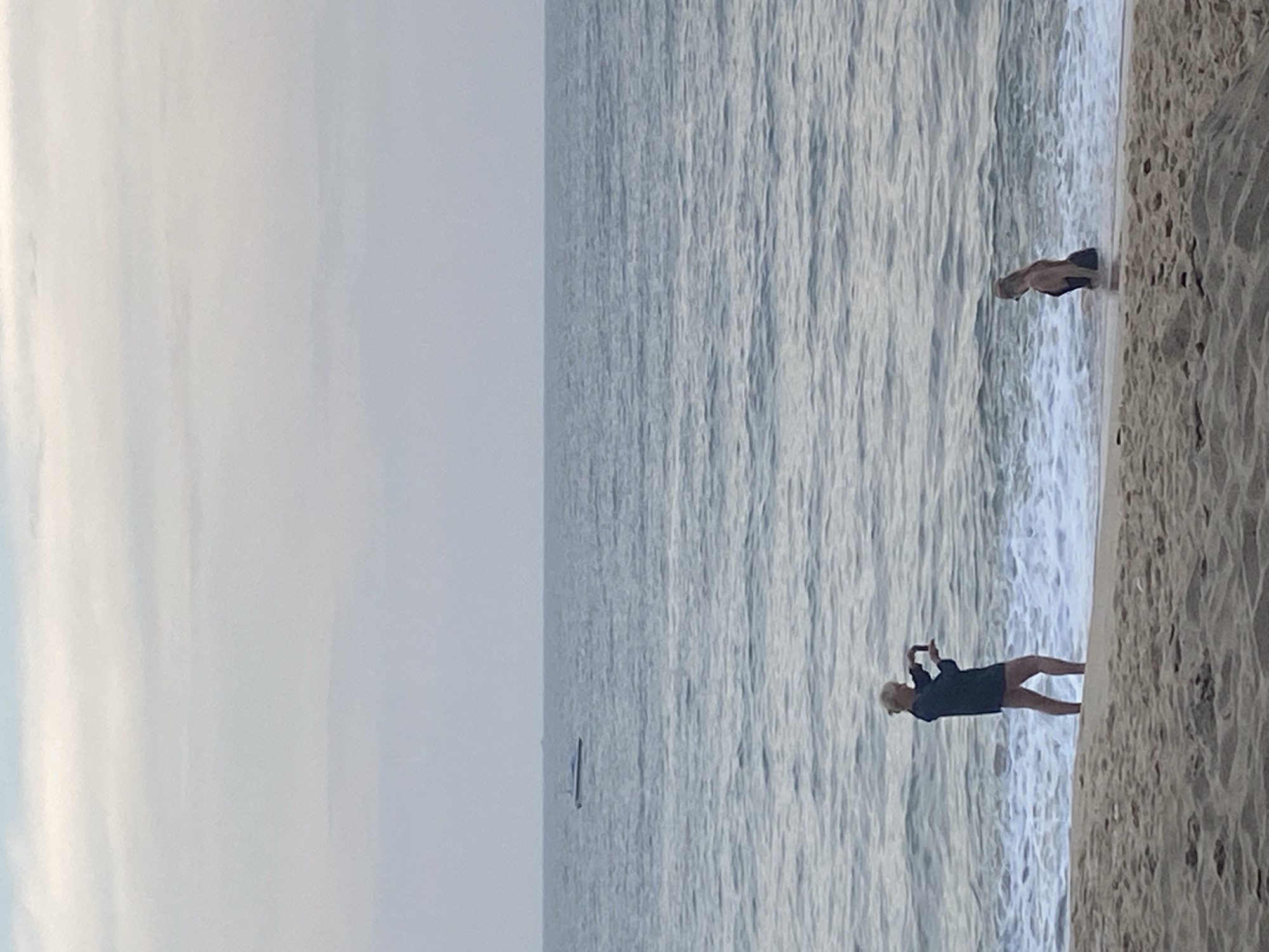An overcast, hazy day at a beach in Mũi Né, Vietnam. The sea is calm with moderate waves rolling onto a sandy shore. In the distance, a small boat is visible on the horizon.
In the foreground, two individuals are present. On the left, a woman stands partially in the surf, facing away from the camera. She is wearing a dark, long-sleeved jacket over dark shorts, and is holding a mobile phone in her hands, appearing to take a photo. Her hair is pulled back in a bun.
To the right of the first woman, another woman is partially submerged in the water. She is wearing a dark swimsuit and has her back to the camera. Her hair is blonde and pulled back. The overall impression is a serene, quiet moment on the beach.
