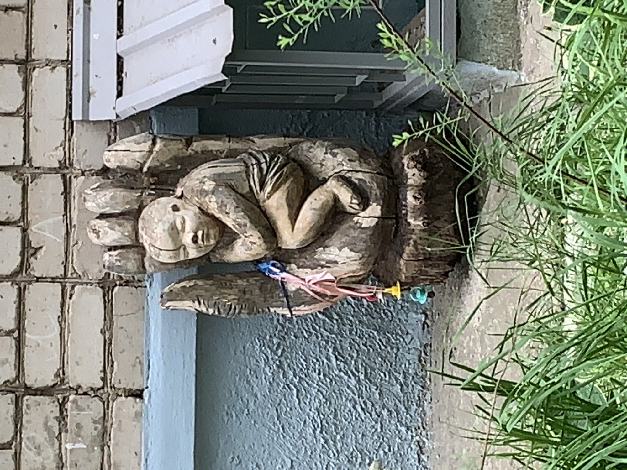 A wooden sculpture of a person lying curled up in a hand is affixed to the exterior wall of a building. There are ribbon-like objects and other items tied to the sculpture, appearing as offerings. The wall is made of bricks, some painted light blue near the sculpture. A white ledge or structure is visible above the sculpture. Green plants partially obscure the right side of the image. The location is likely Chaikovsky, Russia, but there are no identifying Russian elements to guarantee it.