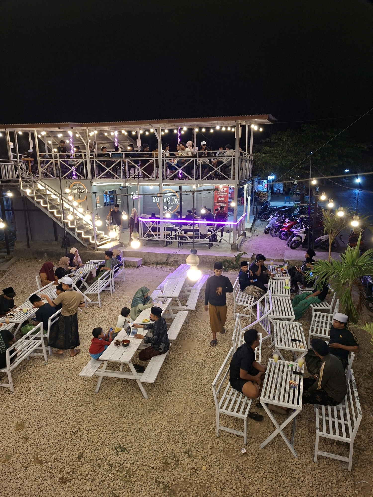 The image depicts an outdoor dining area at night, likely a restaurant or cafe, in Lantek Barat, Indonesia. The establishment features a two-story structure with an open-air balcony on the upper level, illuminated by string lights. On the ground level, several groups of people are seated at white picnic tables situated on a gravel surface. Many patrons are dressed in traditional Islamic attire, including peci caps.
In the foreground, to the left, a group of individuals, including a child, are seated at a table. To the right, another table is occupied by a man working on a laptop. Further to the right, a group of men are engaged in conversation, with drinks and snacks on their table. In the background, motorcycles are parked, suggesting a local clientele. The overall ambiance is casual and social, with the warm glow of the lights creating an inviting atmosphere.