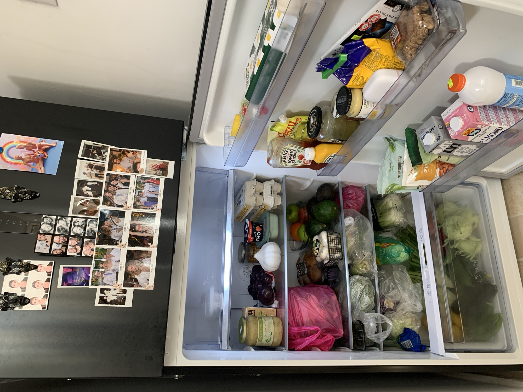 The image shows the interior of a refrigerator. The refrigerator is stocked with various food items, including fruits, vegetables, condiments, dairy products, and snacks. The door of the refrigerator has photographs and magnets attached to it. Labels on some of the food items are in Hebrew, suggesting the location is likely in Israel, possibly Tel-Aviv.