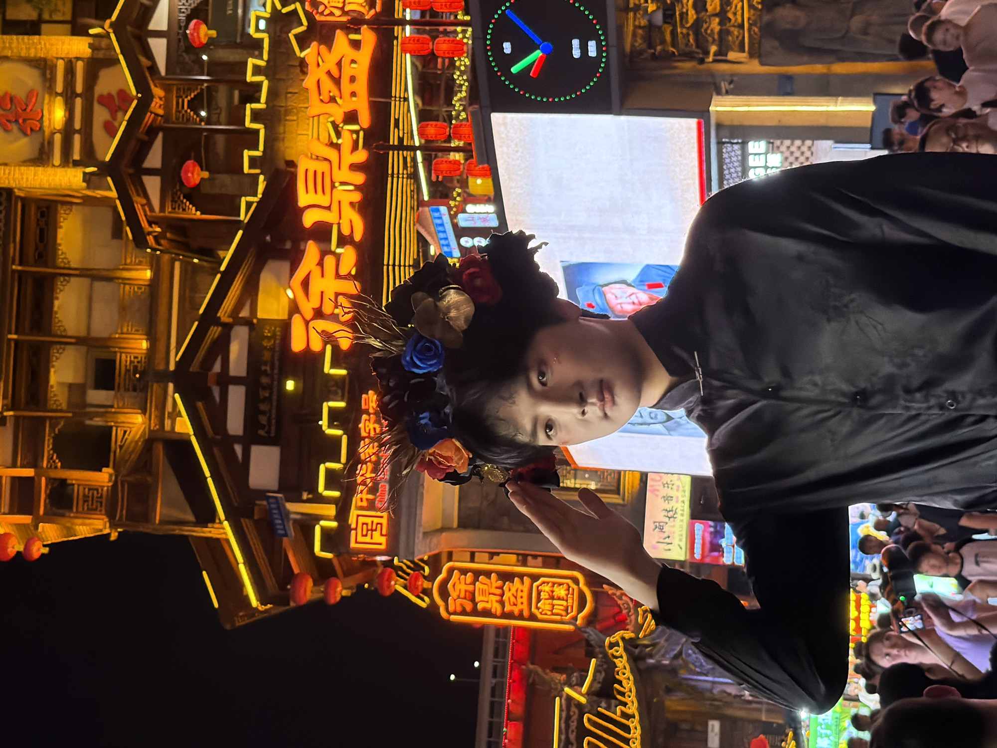 This media file, captured in Yuzhong District, China, presents a vibrant nighttime scene at what appears to be a bustling street market or a traditional entertainment district. The focal point is a young person wearing a decorative headpiece adorned with artificial flowers. They are looking towards the camera, with one hand raised in a gesture that could be a greeting or a pose.
The background is dominated by traditional Chinese architecture illuminated by a profusion of neon signs and lanterns. These lights cast a warm glow on the wooden structures and intricate facades, creating a festive and lively atmosphere. The signs themselves feature Chinese characters, with some legible text including "中华老字号" (China's Time-Honored Brand), "徐鼎盛" (Xu Dingsheng), "渝味" (Yu Flavor), and "川菜" (Sichuan Cuisine), indicating the presence of established food establishments. A large clock with a digital display is also visible in the upper right section.
The presence of a crowd of people, some holding up phones, suggests that this is a popular location, likely a tourist attraction or a place for evening entertainment. The overall mood is energetic and visually rich, with the bright lights and traditional aesthetics contributing to a sense of cultural immersion. The time of day is clearly night, indicated by the darkness of the sky and the prominence of artificial lighting. There are no obvious weather cues.