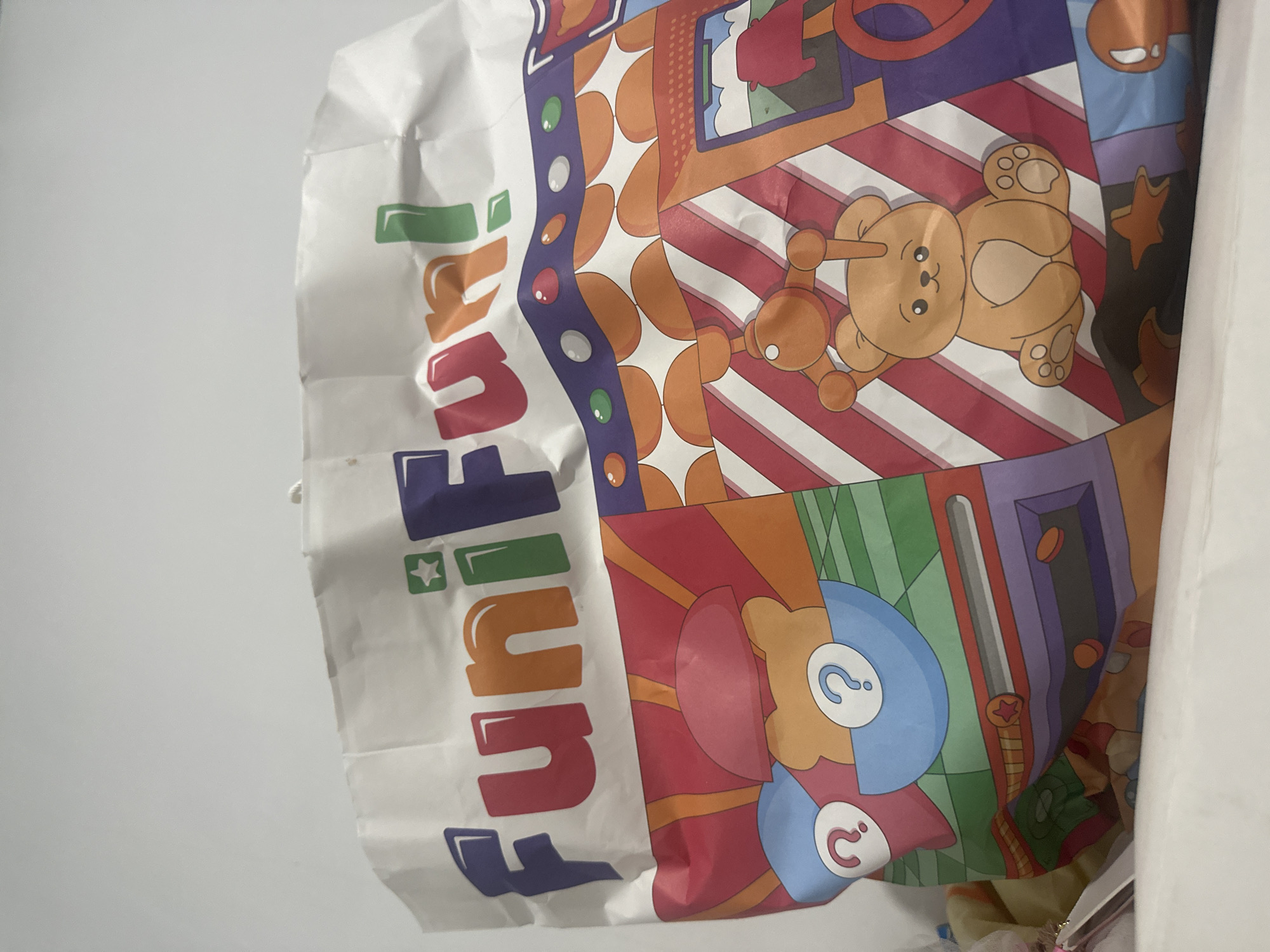The image shows a close-up of a white paper shopping bag with colorful cartoon graphics. The bag is branded with the words "FunFun!" in large, stylized letters across the top, with a star above the first "i" and an exclamation mark after the second "n". The bottom portion of the bag is decorated with various playful imagery, including a smiling cartoon teddy bear, question marks within circles, geometric patterns, and what appears to be a scene from a carnival or arcade game. The bag is slightly crumpled, and its top edge is visible against a plain white background. There are no people or visible elements that suggest a specific location within the city of Binjai, Indonesia.