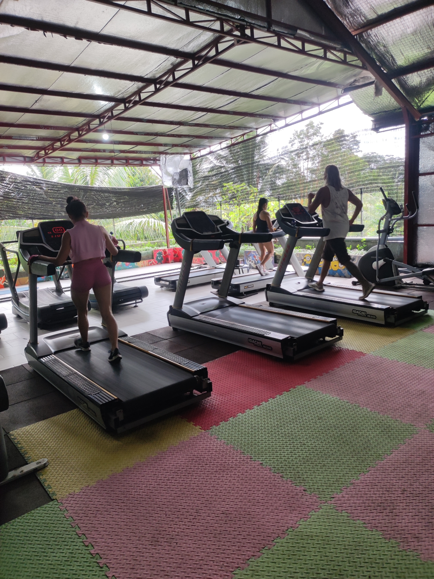 The image depicts a fitness gym in Kedewatan, Indonesia. Three individuals are using treadmills. One woman is wearing pink athletic wear, another woman is wearing a black athletic outfit, and a man is wearing a white tank top with dark shorts. The gym has a metal frame roof, open sides, and a view of lush green trees outside. The floor is covered with multi-colored interlocking foam mats. Various exercise machines are present in the background. The lighting suggests it is daytime.