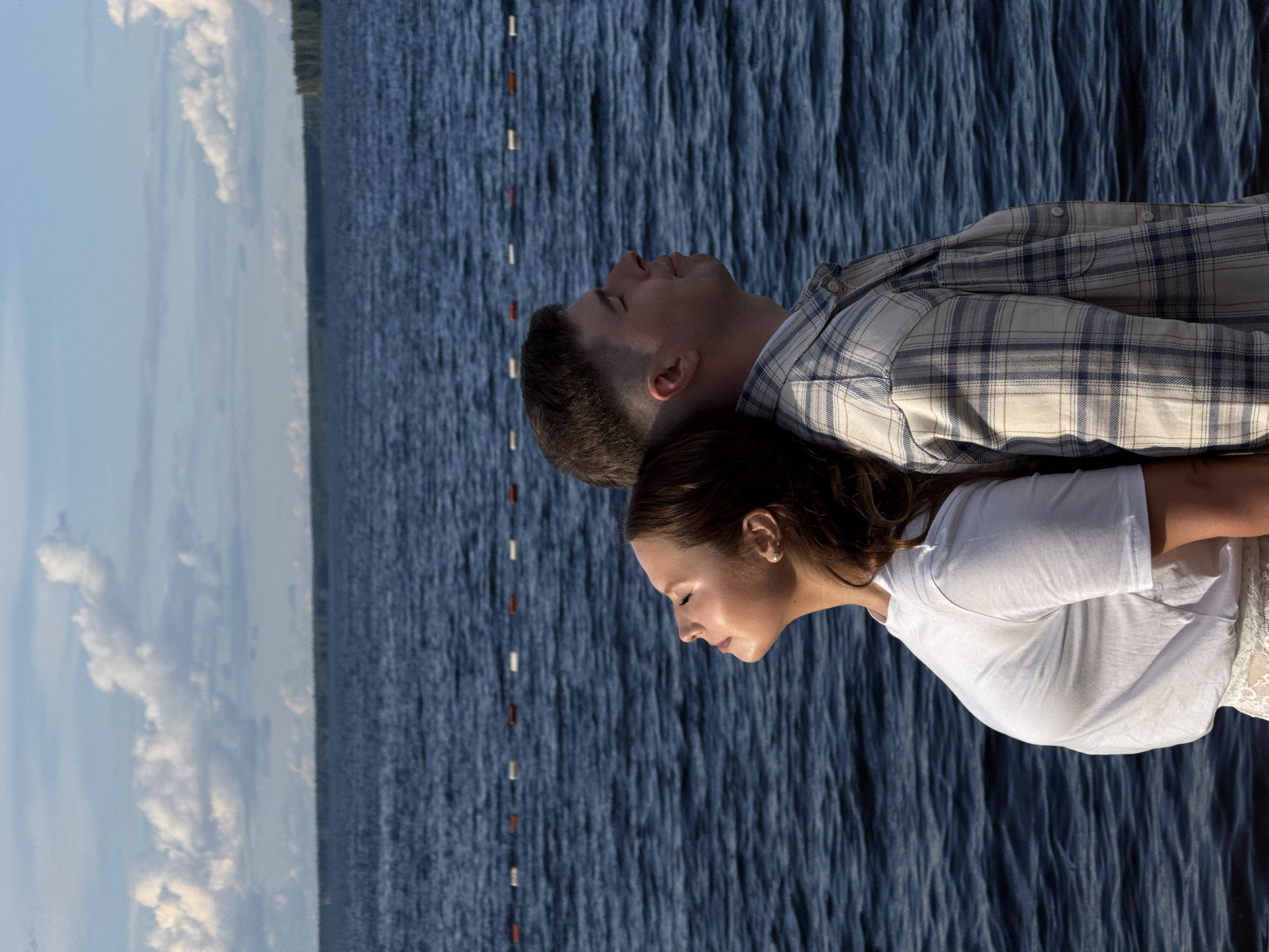 A couple stands next to water under a cloudy sky. The man, with short brown hair, wears a light plaid shirt and tilts his head back. The woman, with long brown hair, wears a white t-shirt and keeps her head down. In the background, there is a body of water with buoys. This image was taken in Chaikovsky, Russia.