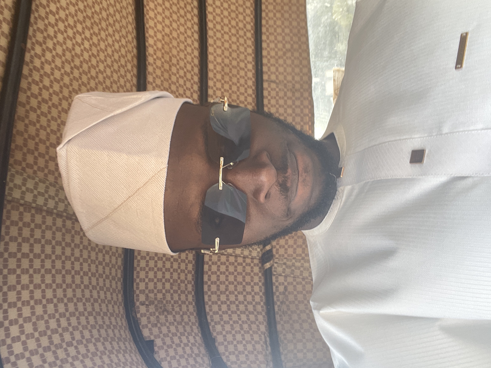 A man is depicted wearing a traditional cream-colored attire with a matching cap and sunglasses. He is positioned against a patterned brown and cream backdrop, suggestive of an interior setting. The photograph appears to be taken indoors, possibly inside a building. Due to the lack of landmarks or specific contextual details, it is not possible to conclusively determine if the location is in Jos, Nigeria.