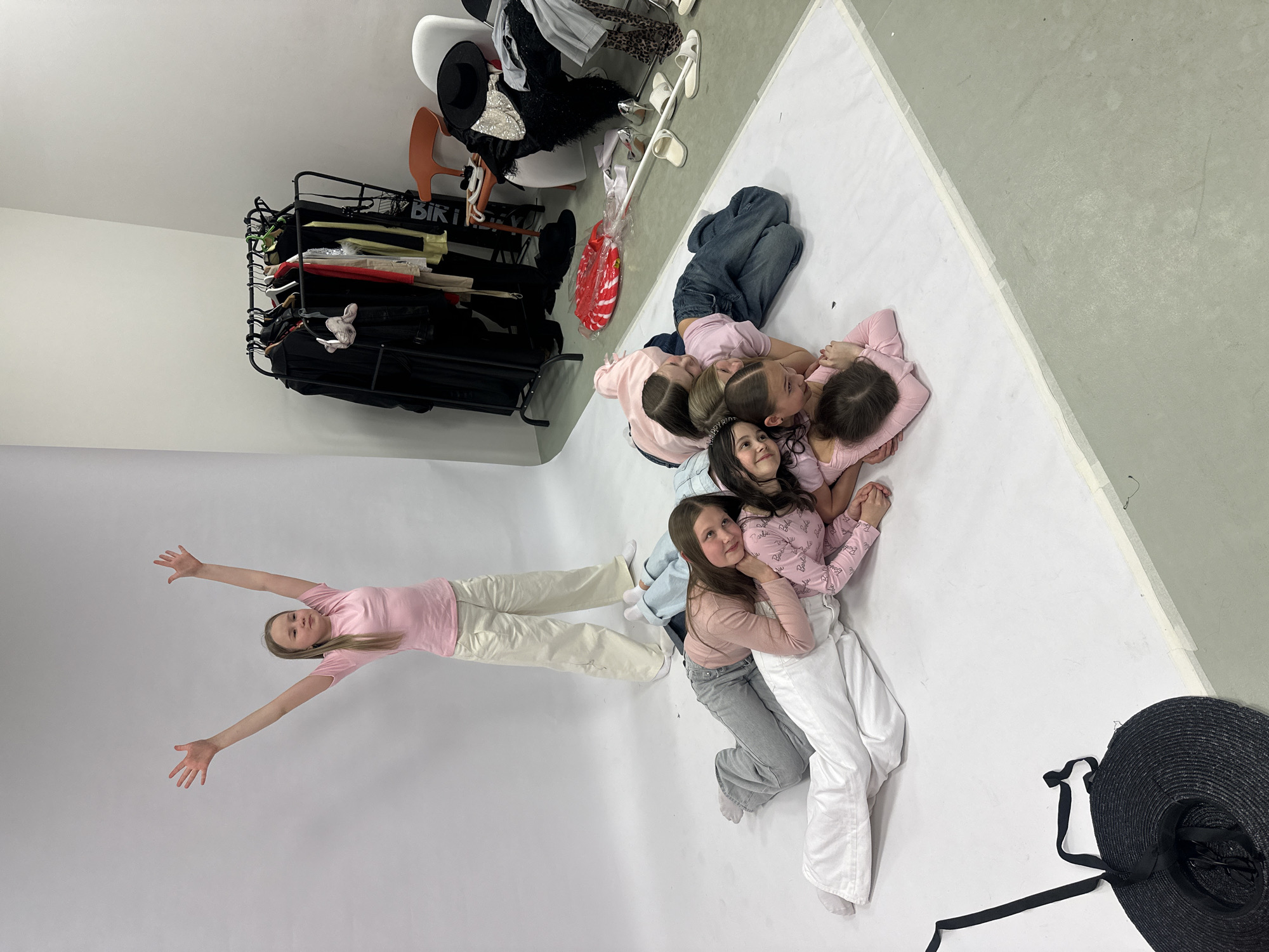 A photograph taken indoors shows six young females lying on a large white backdrop. The composition is from an overhead angle.
One girl, wearing a pink shirt and light-colored pants, is positioned with her head near the left edge of the frame and her arms outstretched. Her body forms a horizontal line. The other five girls are lying in a pile, with their bodies running along a vertical axis, towards the right edge of the frame. They are wearing a combination of pink shirts and light-colored pants.
In the upper left corner of the photograph, there is a clothing rack with a variety of items, including black clothing, and what appears to be a sign partially visible. To the right of the clothing rack is a collection of objects, including an orange chair, and a white chair with additional clothing.
A large black hat is in the lower right corner of the photograph.
The location cannot be determined to be Votkinsk, Russia based on the visual information.