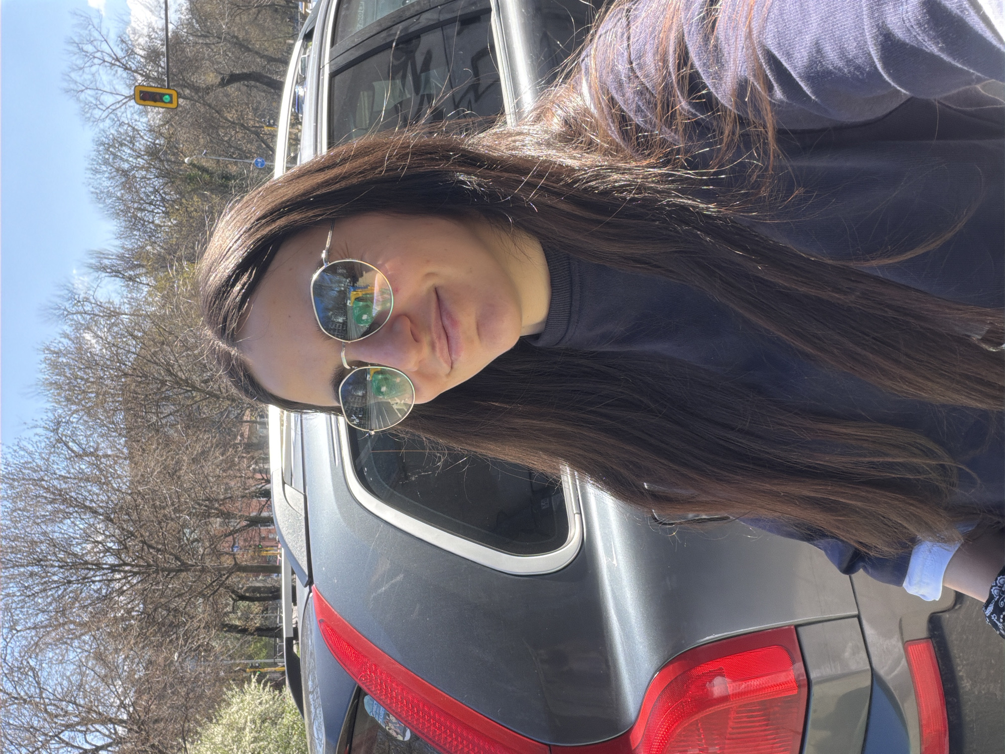 A woman with long, brown hair and sunglasses stands next to the rear of a gray Volvo SUV on a city street. The background reveals trees with bare branches, indicating a season like early spring or late fall. A traffic light is visible in the upper left corner of the frame. Reflections in the sunglasses suggest an urban environment, with buildings and street activity. The location is in Sofia, Bulgaria.