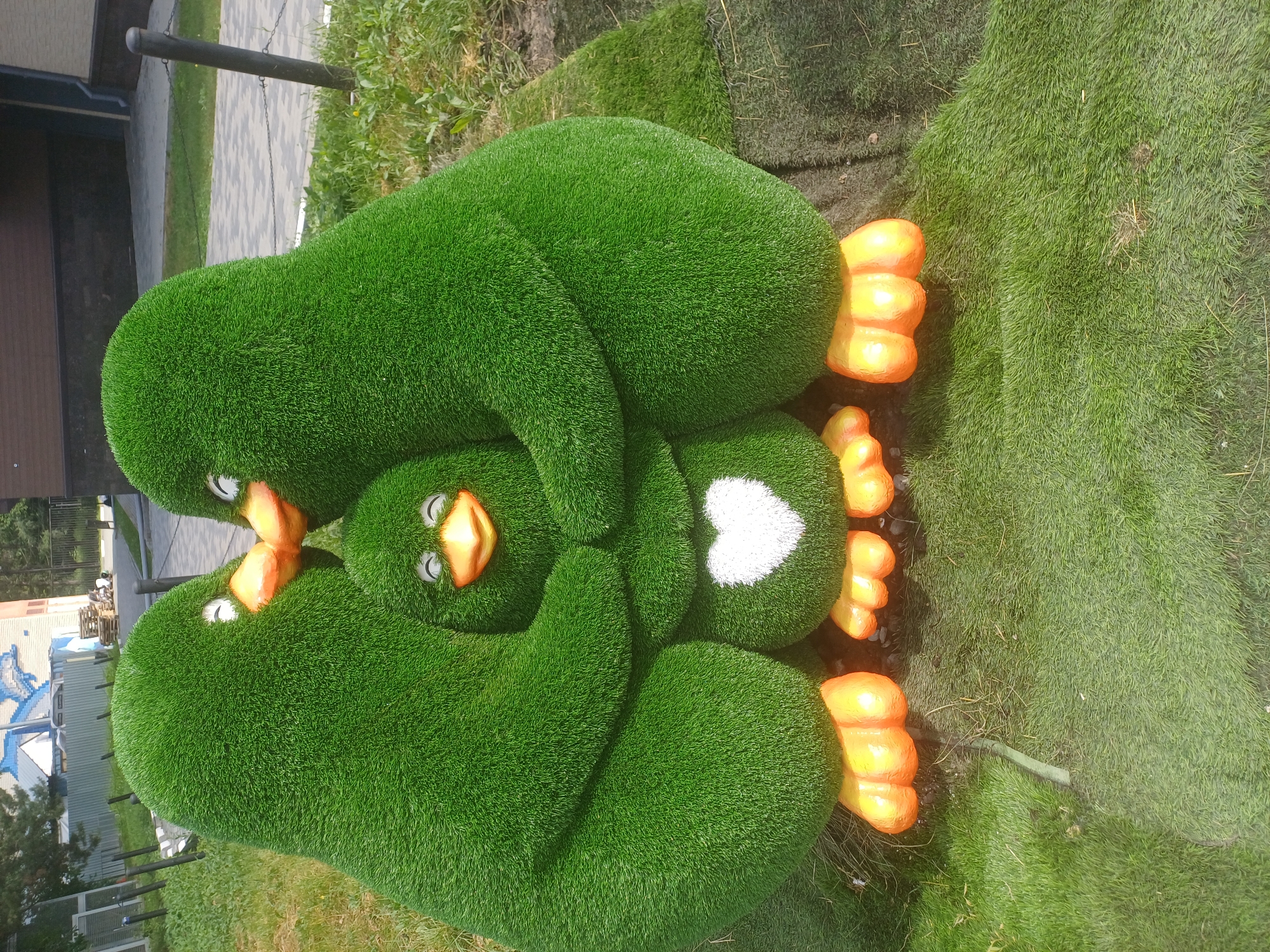 In Novosibirsk, Russia, a grassy sculpture of three penguins is displayed outdoors. The two larger penguins embrace a smaller penguin, which has a white heart design on its chest. All three penguins are green and have orange feet and beaks. The sculpture is situated on a patch of artificial grass surrounded by natural grass and a paved walkway. In the background, a building with a blue roof and a fence can be seen. The image appears to be taken on a sunny day.