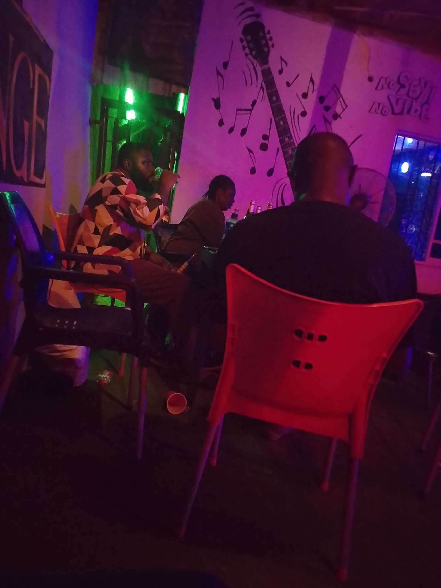The image shows an indoor scene in what appears to be a bar or lounge, possibly in Yelwa, Nigeria. Three people are visible. One man is sitting in the foreground with his back to the camera, wearing a black shirt and sitting in a red plastic chair. Two other men are sitting at a bar or table in the background, one wearing a colorful geometric patterned shirt and the other wearing a brown shirt. A wall behind them is decorated with musical notes and an image of a guitar. The lighting is dark and includes a mix of purple, blue, and green lights, creating a vibrant yet dim atmosphere. There are plastic chairs in the foreground and a cup on the floor.