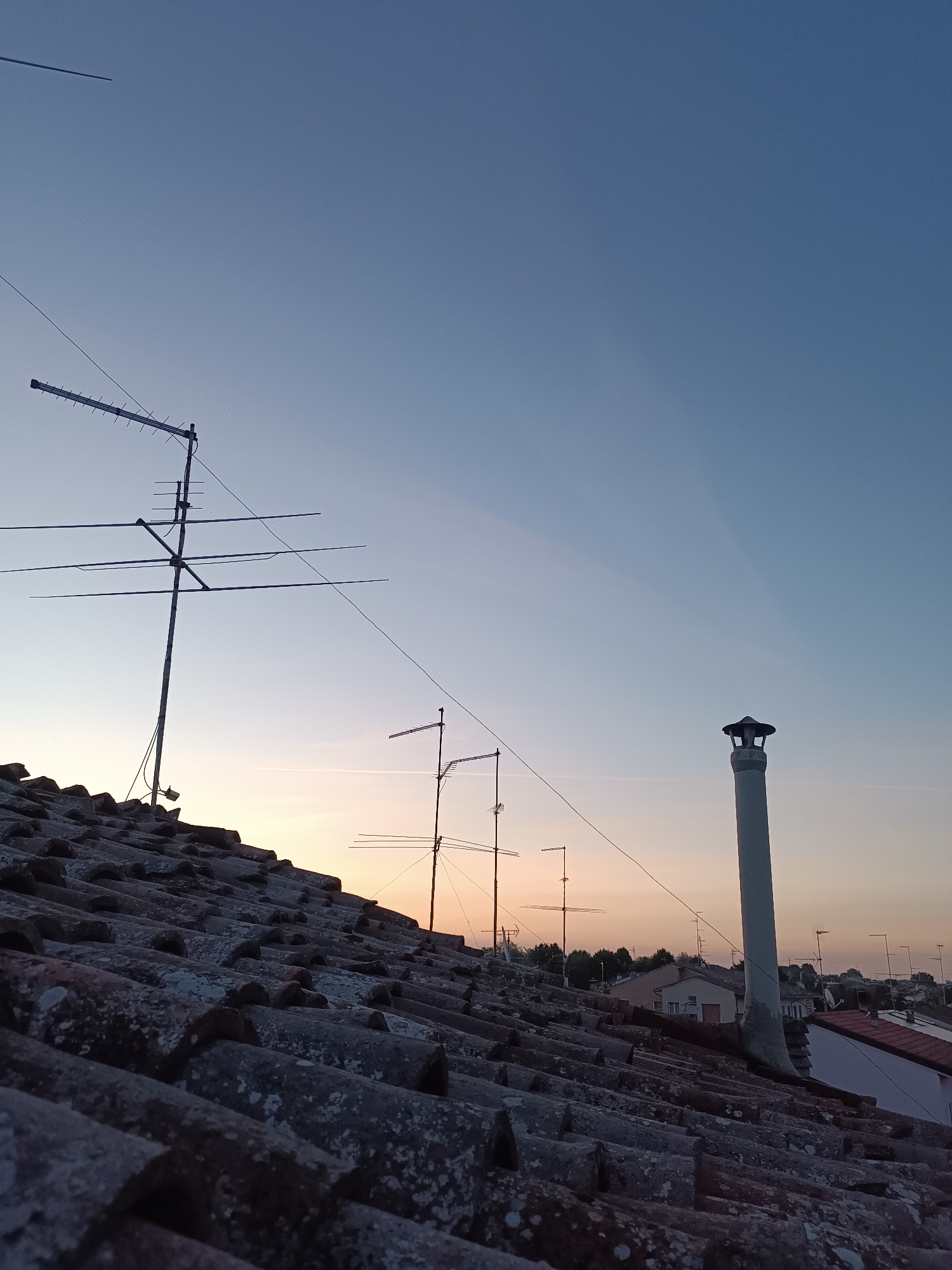 The image shows a rooftop view in Viadana, Italy, at twilight. The foreground is dominated by a close-up of weathered, red clay roof tiles, speckled with lichen. In the mid-ground, several television antennas are visible, silhouetted against a gradient sky that transitions from pale blue at the top to a warm orange near the horizon. A tall, white chimney with a cap stands prominently to the right. In the distance, more rooftops and the silhouettes of trees and buildings are discernible, indicating an urban or suburban setting. The sky is clear, with faint contrails or atmospheric streaks visible. There are no people or visible actions taking place. The overall impression is one of a quiet, residential area at the end of the day.