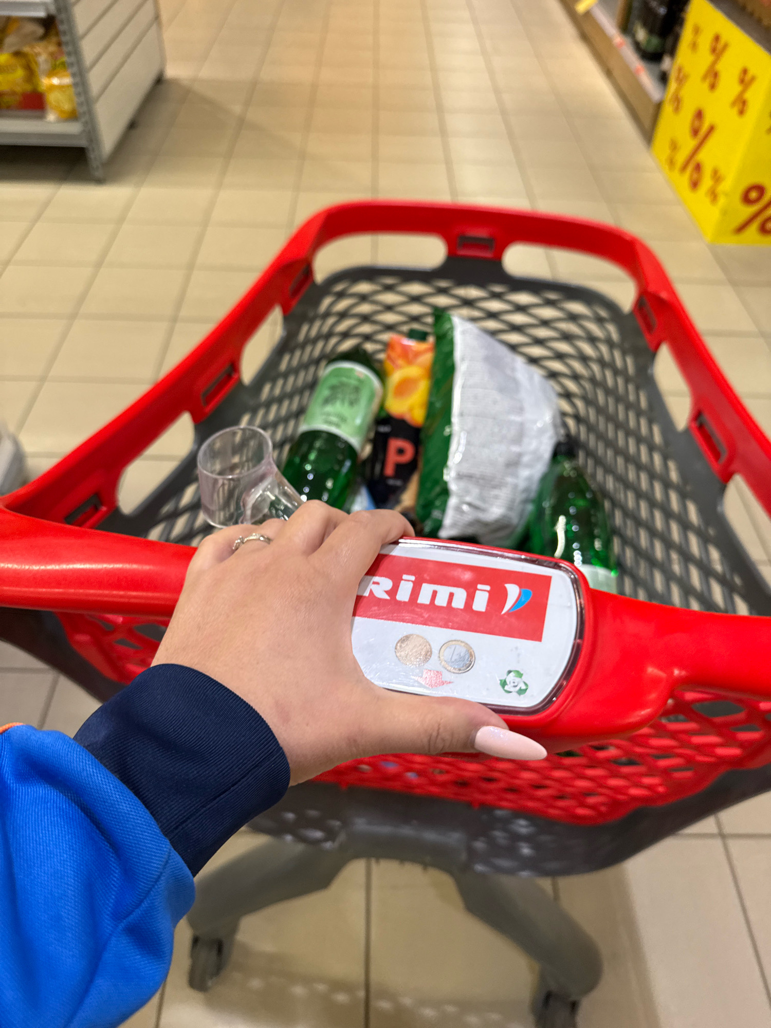 A first-person perspective shot of a shopping cart in a Rimi supermarket in Tallinn, Estonia. The cart is red and black and contains several items, including bottles of green liquid, a bag of chips, and a white bag. A hand with a ring on its finger and light pink nails is gripping the handle of the cart. The floor is tiled with beige and brown squares. In the background, there are shelves stocked with various products, and a yellow sign with red percentage symbols is visible on the right.