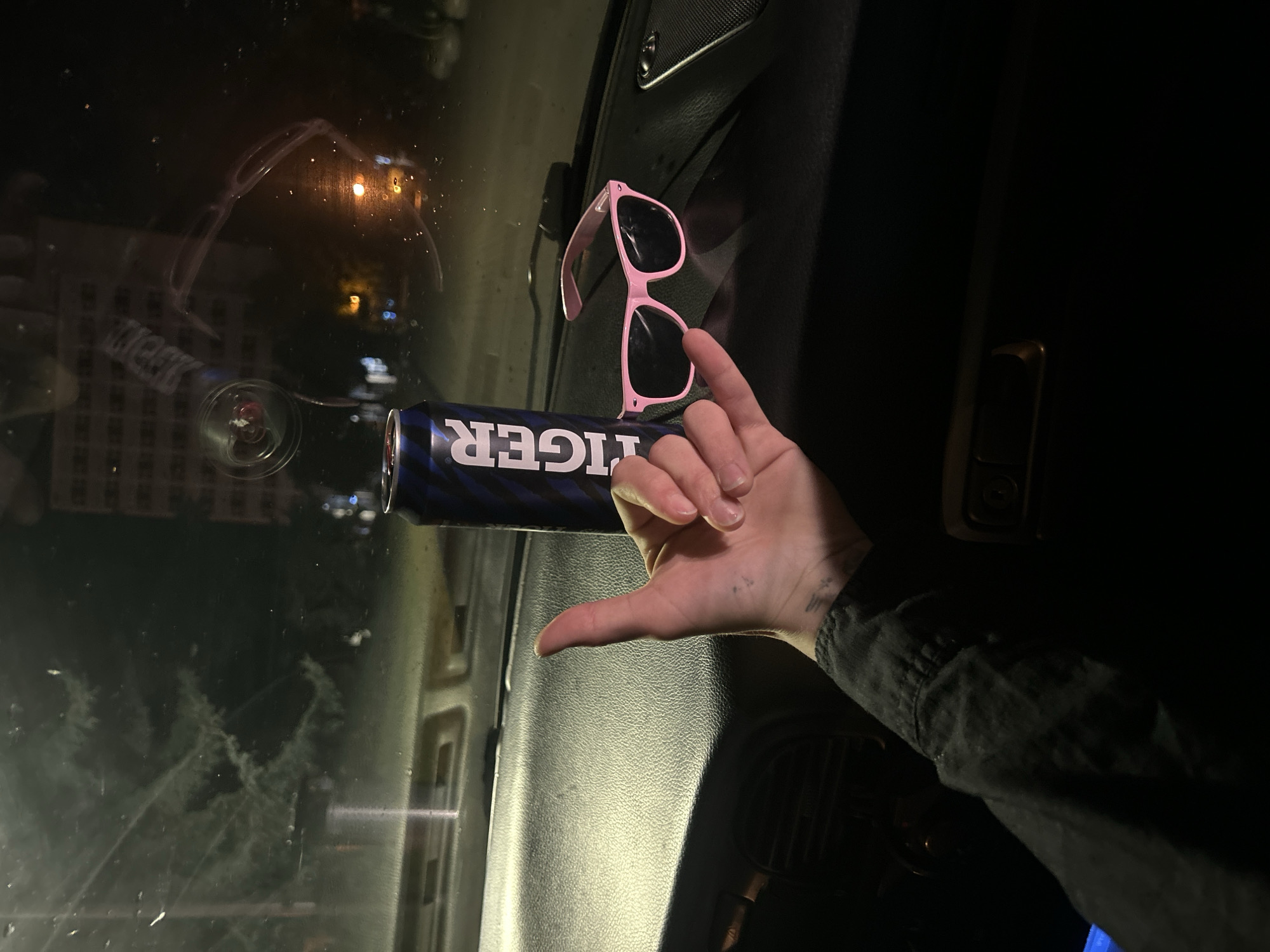 An overhead shot shows a hand holding up a "shaka" sign, with a can of Tiger beer and a pair of pink sunglasses resting on the dashboard of a vehicle. The scene is lit by the dim interior lights of the car, and the exterior view through the windshield is dark, suggesting nighttime. The background reveals a city scene with buildings and streetlights, possibly indicating an urban environment. The car's interior shows the textured dashboard typical of a vehicle. The overall impression is a casual moment captured inside a car at night. There is no specific information in the image that indicates the location to be Siedlce, Poland.