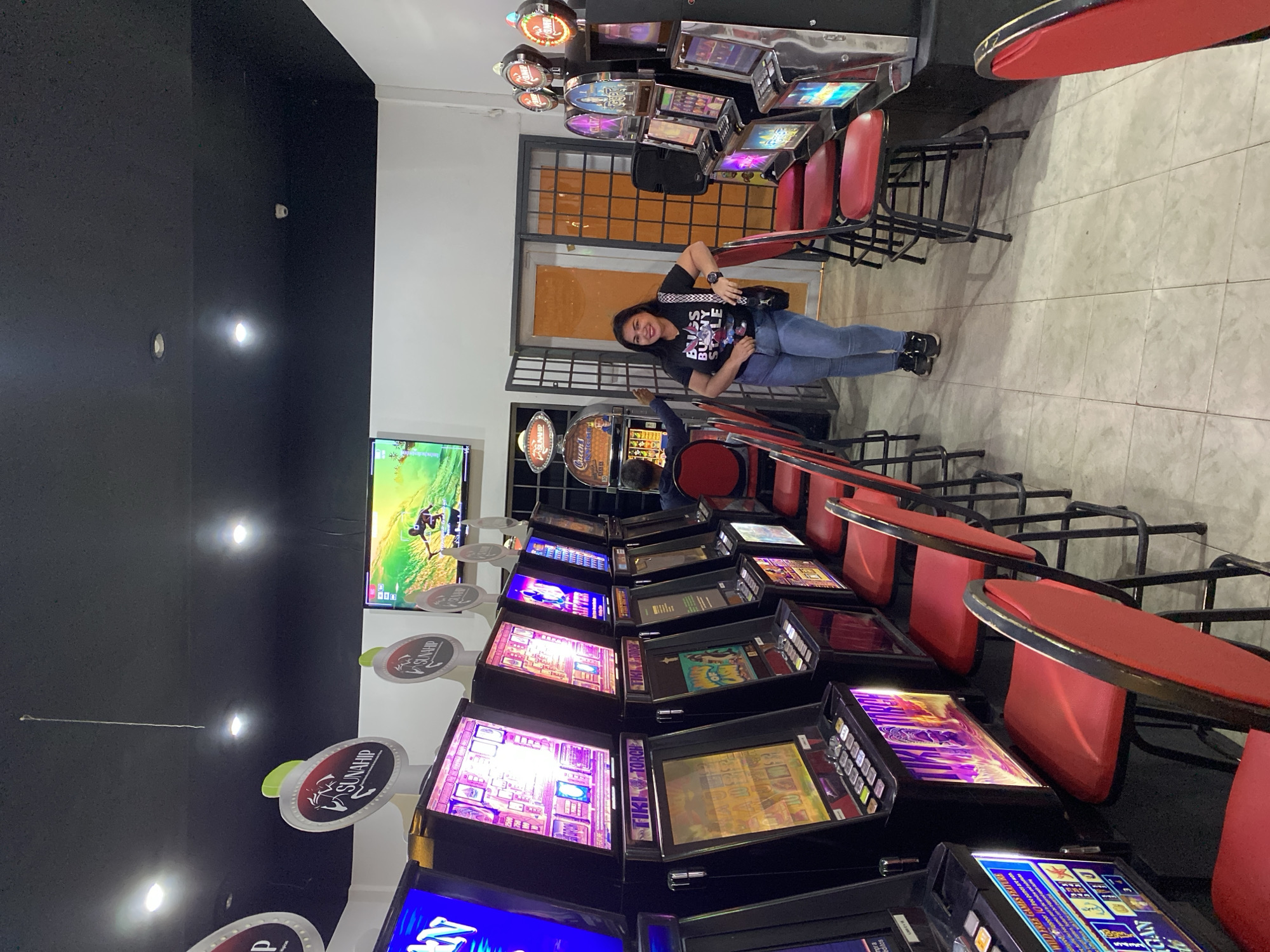 The image shows the interior of a casino in Juan Griego, Venezuela. The walls are painted black. There are rows of slot machines with red chairs in front of them. A flat screen TV is mounted on the wall in the background. A woman is standing near the entrance. She is wearing a Bugs Bunny t-shirt, jeans, and sneakers. She has a black purse slung across her body. The floor is tiled. There are ceiling lights on the black ceiling.