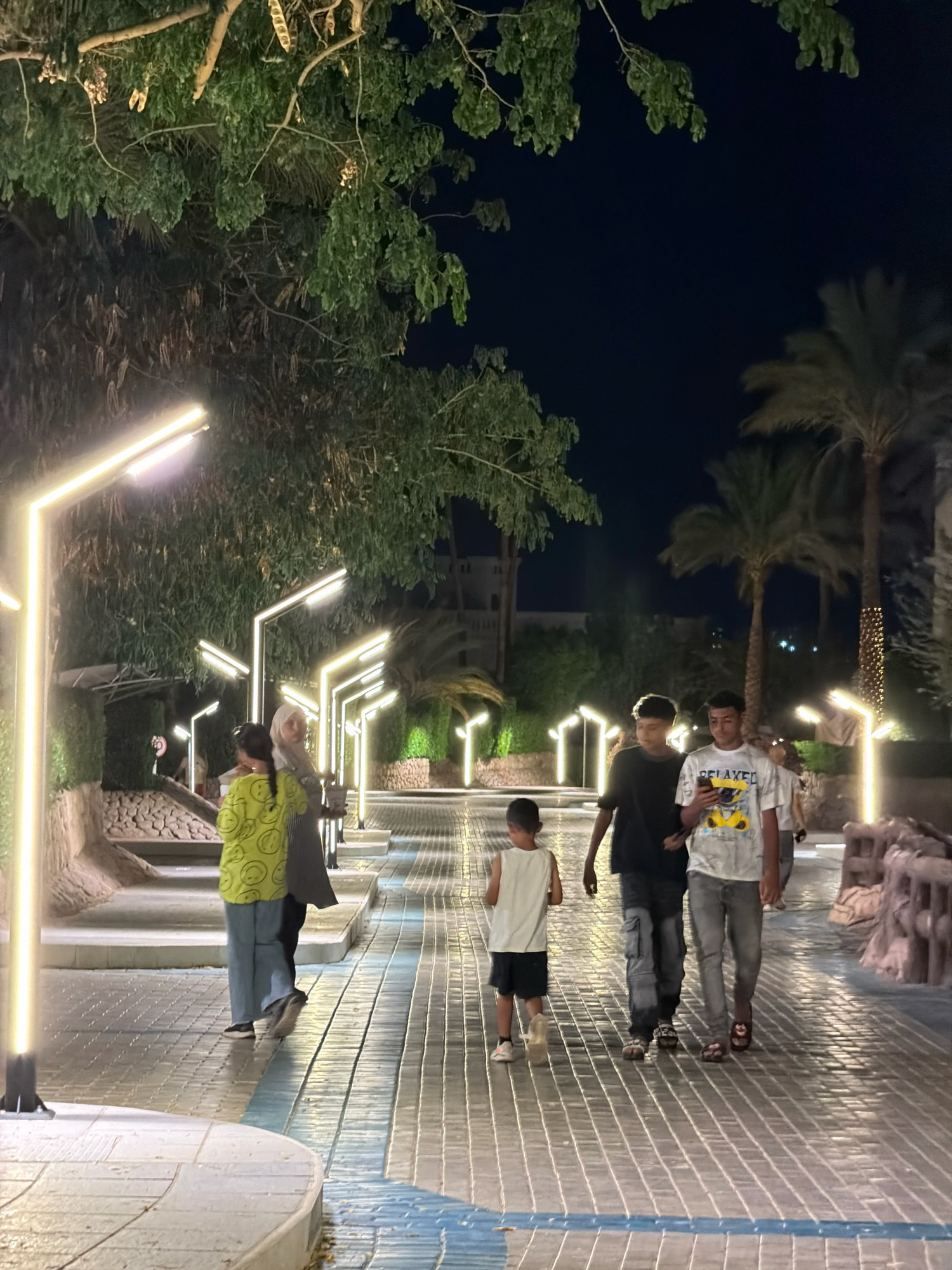 A group of people walk along a illuminated pathway at night in Sharm El Sheikh, Egypt. The pathway is paved with textured tiles and lined with modern, angular lampposts emitting a warm yellow light. Lush green trees and palm trees frame the scene, casting shadows on the ground. In the foreground, two young boys walk side-by-side, one in a black t-shirt and cargo pants, the other in a white t-shirt with a graphic print and grey jeans. Behind them, a younger boy in a white tank top and dark shorts walks alone. Further down the path, a woman wearing a headscarf and a patterned green shirt walks with another person. The background features architectural structures and more distant lighting, indicating an urban or resort setting.