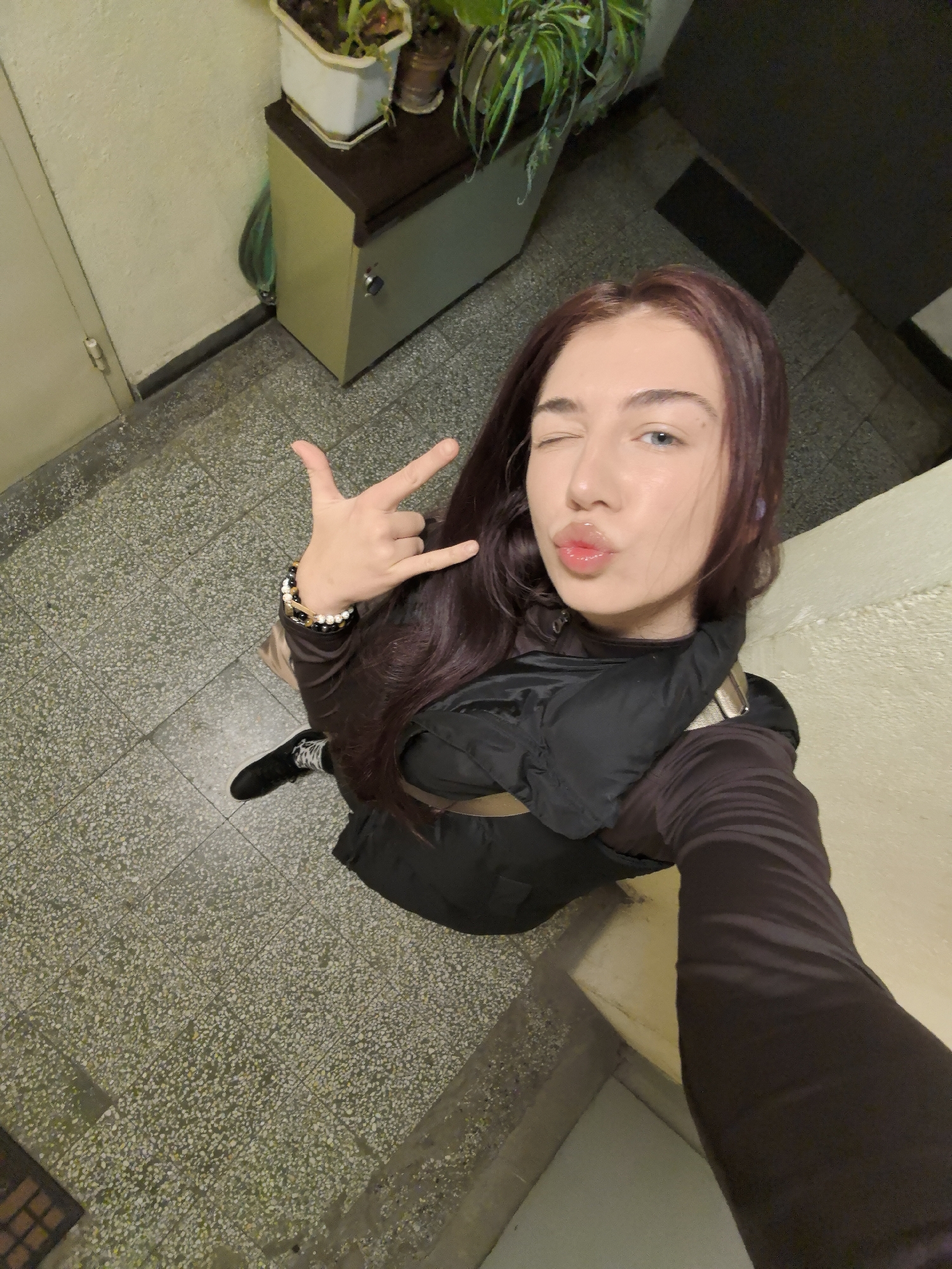 A young woman poses indoors, likely in an apartment building hallway, based on the tile floor, door, and electrical closet/cabinet with potted plants on top. She is wearing a dark jacket, a long-sleeved shirt, and has on bracelets. She is holding her arm out, taking a selfie, making a rock-on hand gesture, winking, and pouting her lips. The location in Stara Zagora, Bulgaria, cannot be determined from this photo.