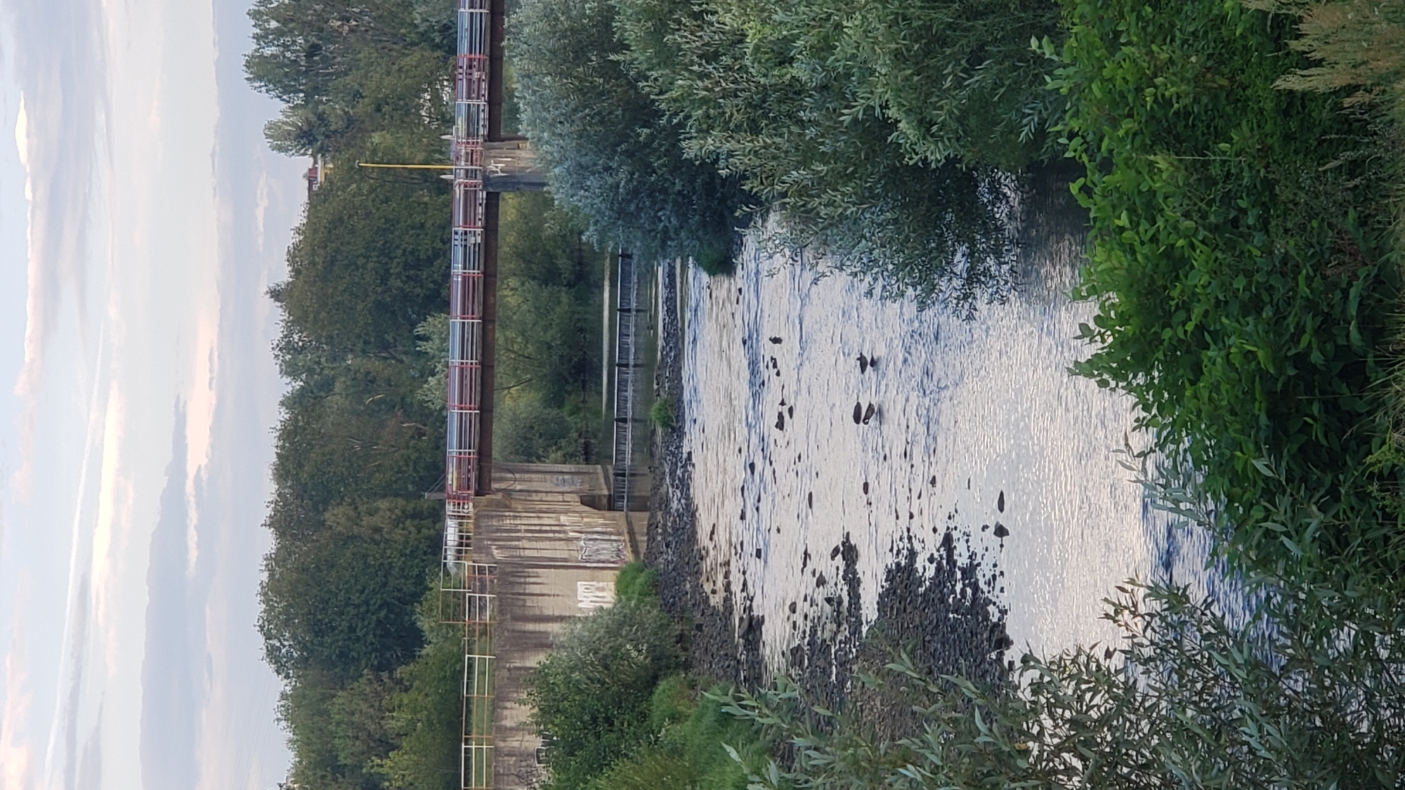 The image shows a river flowing under a bridge with a dam. The bridge has a multi-colored railing and concrete pillars. The dam is made of concrete and has a series of small waterfalls. The riverbanks are lined with trees and bushes. There are several ducks in the water. The sky is cloudy. The scene is likely located in the city of Prešov, Slovakia, as indicated by the context.